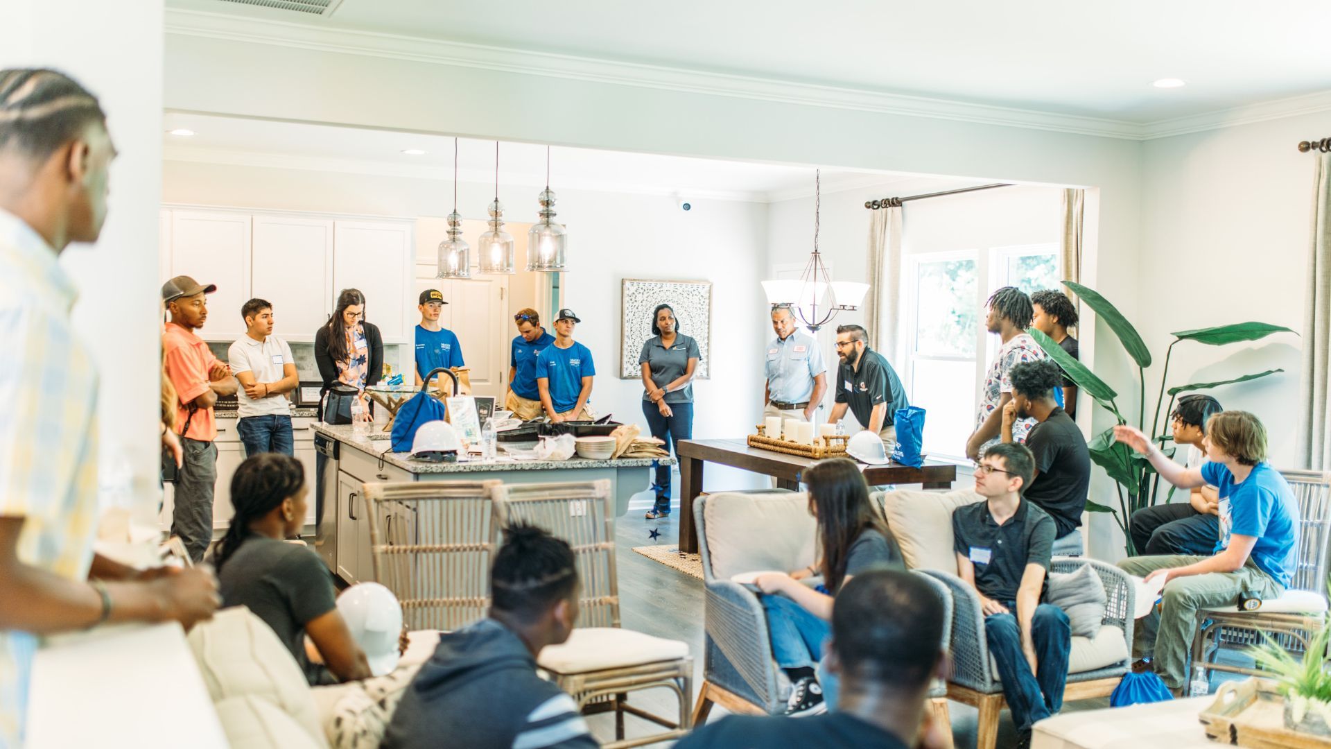 A group of people sitting around a living and dining room, listening to a man speak.