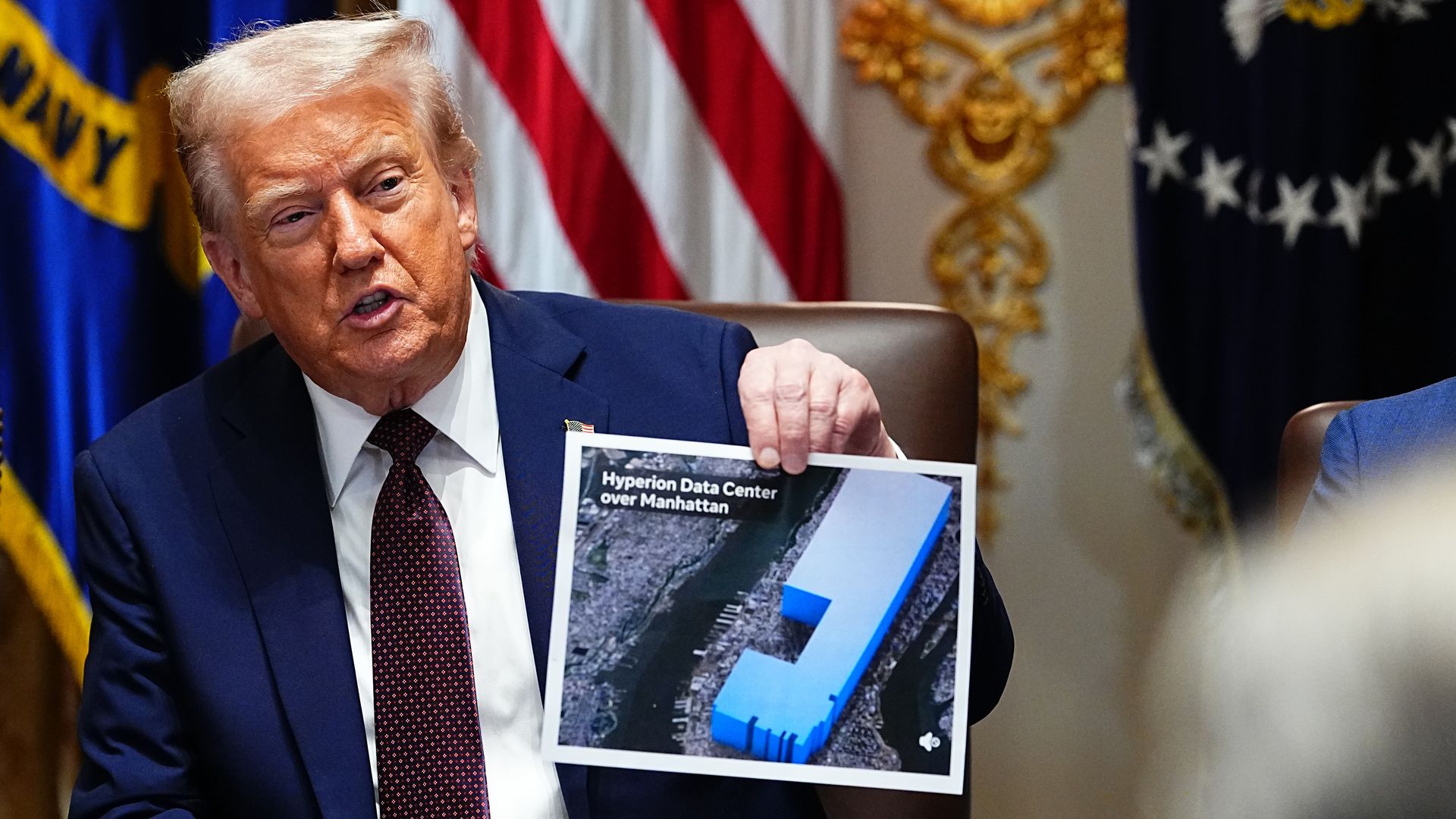 A man in a suit holds a paper with a blue 3D model labeled "Hyperion Data Center over Manhattan" in a room with American flags and ornate gold decorations.