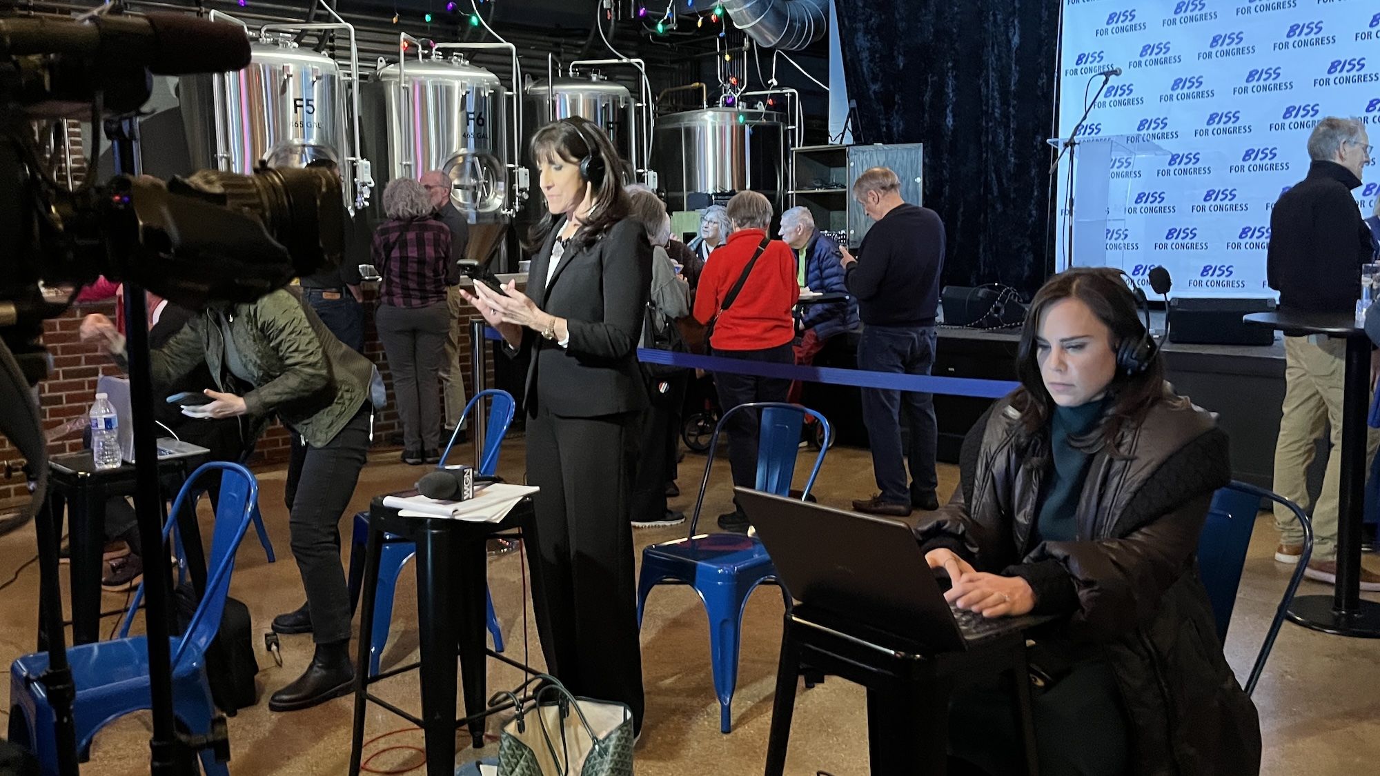 Press area near a stage with a "BISS FOR CONGRESS" backdrop. A suited woman with headset checks her phone; another woman types on a laptop as people queue behind a blue barrier.