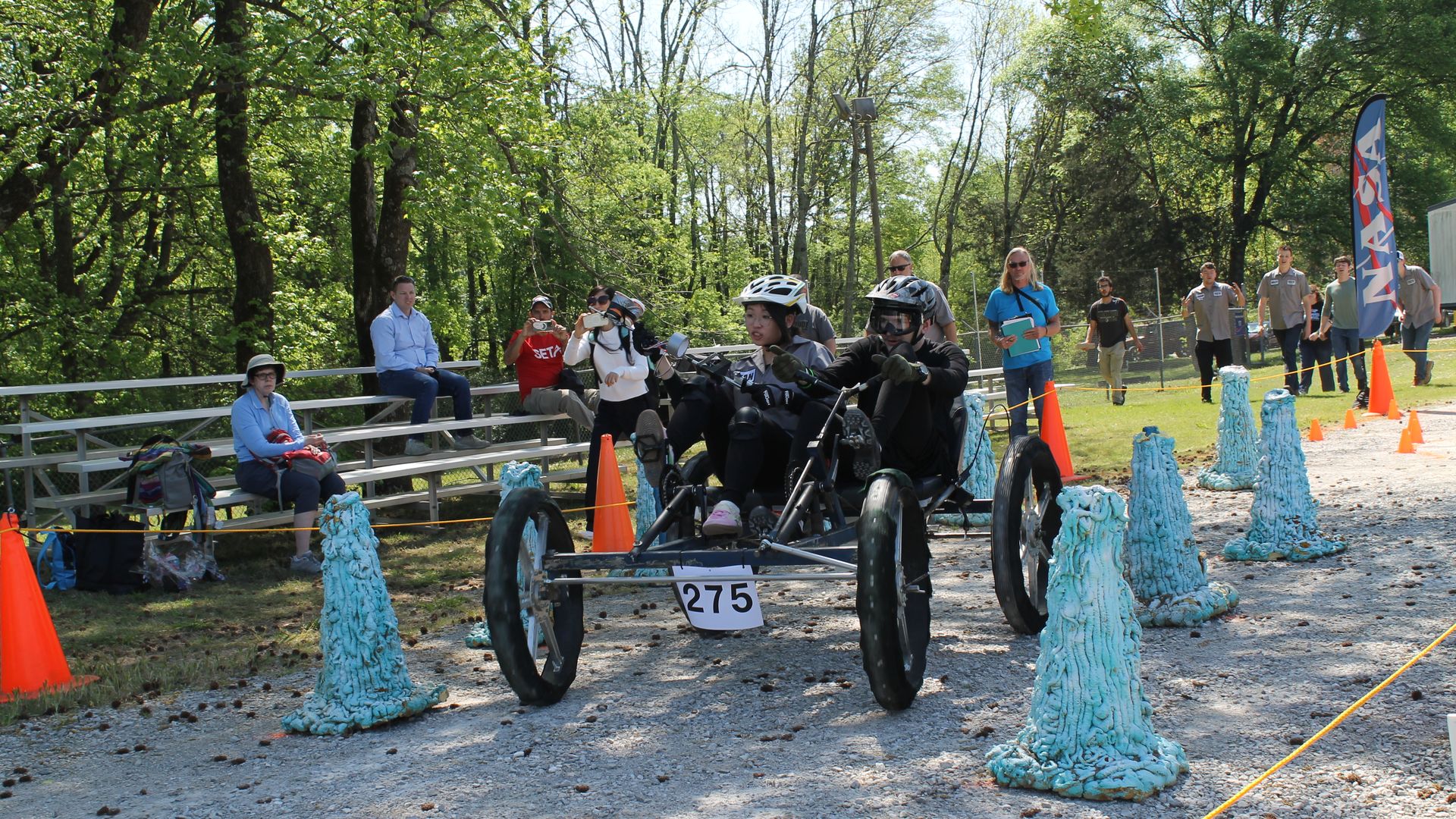 Two helmeted riders steer a blue, three-wheeled cart numbered 275 along a gravel track, with spectators on bleachers and photographers beside orange cones and blue foam obstacles at a race event.