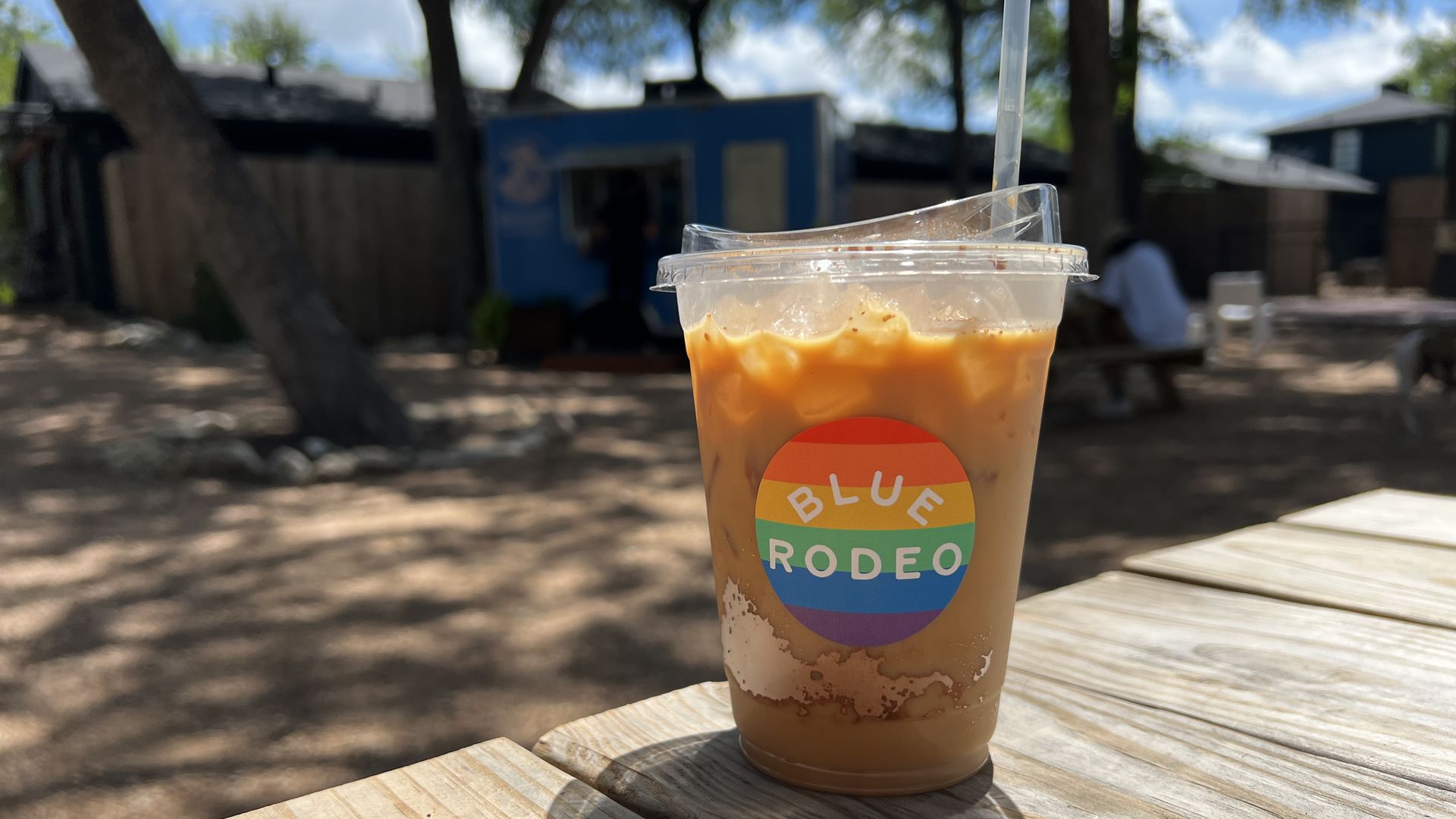 Close-up of iced coffee in a clear cup with lid and straw on a wooden picnic table. Rainbow label reads BLUE RODEO. A sunny outdoor scene with trees and a blue building in the blurred background.