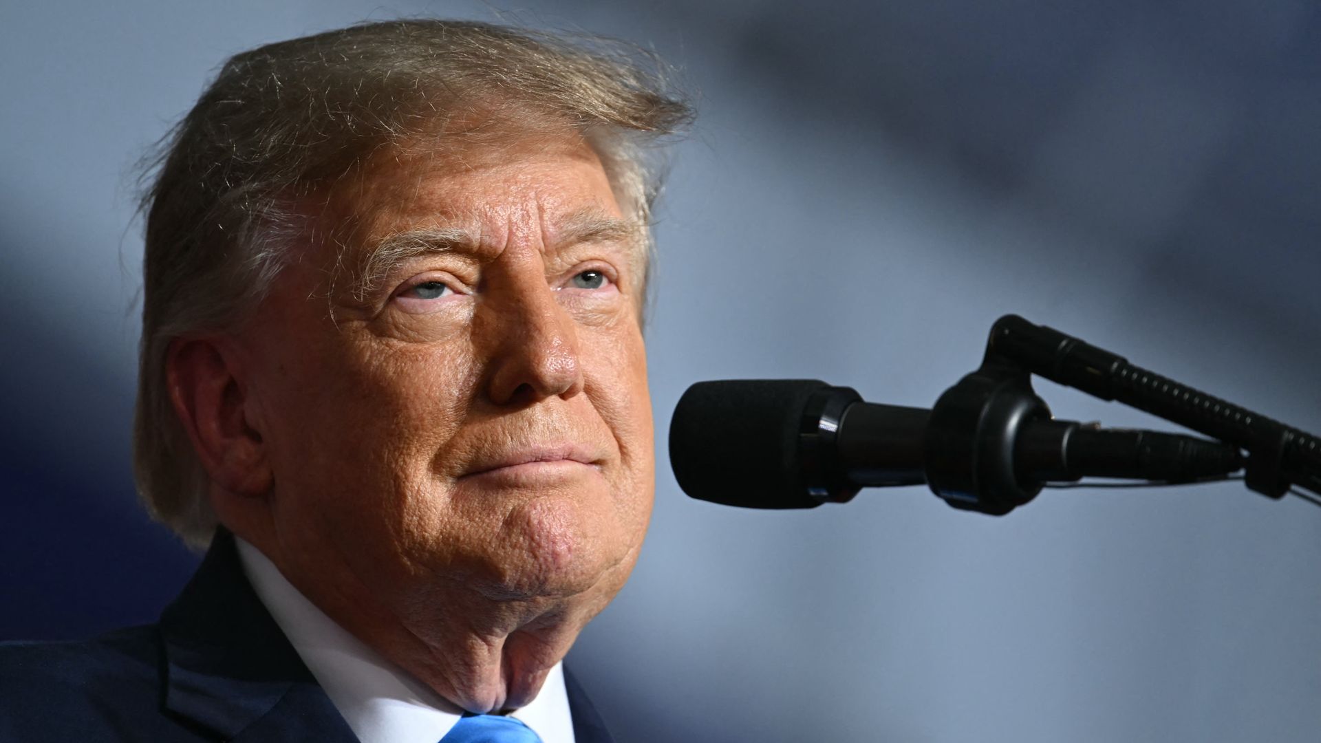 Close-up of President Trump, an older man with gray hair in a dark suit and blue tie, speaking into a microphone. Only part of his face is visible against a blurred blue-gray background.