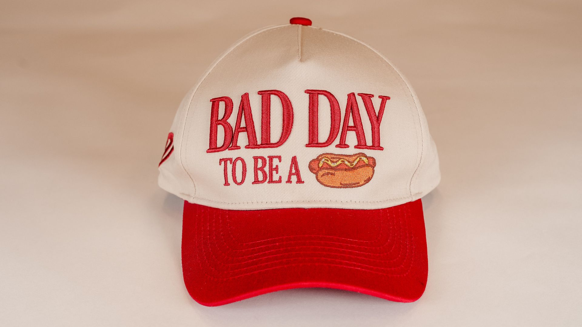 A white cap with a red brim, red embroidered text reading BAD DAY TO BE A and a small hot dog graphic, set against a pale beige background.