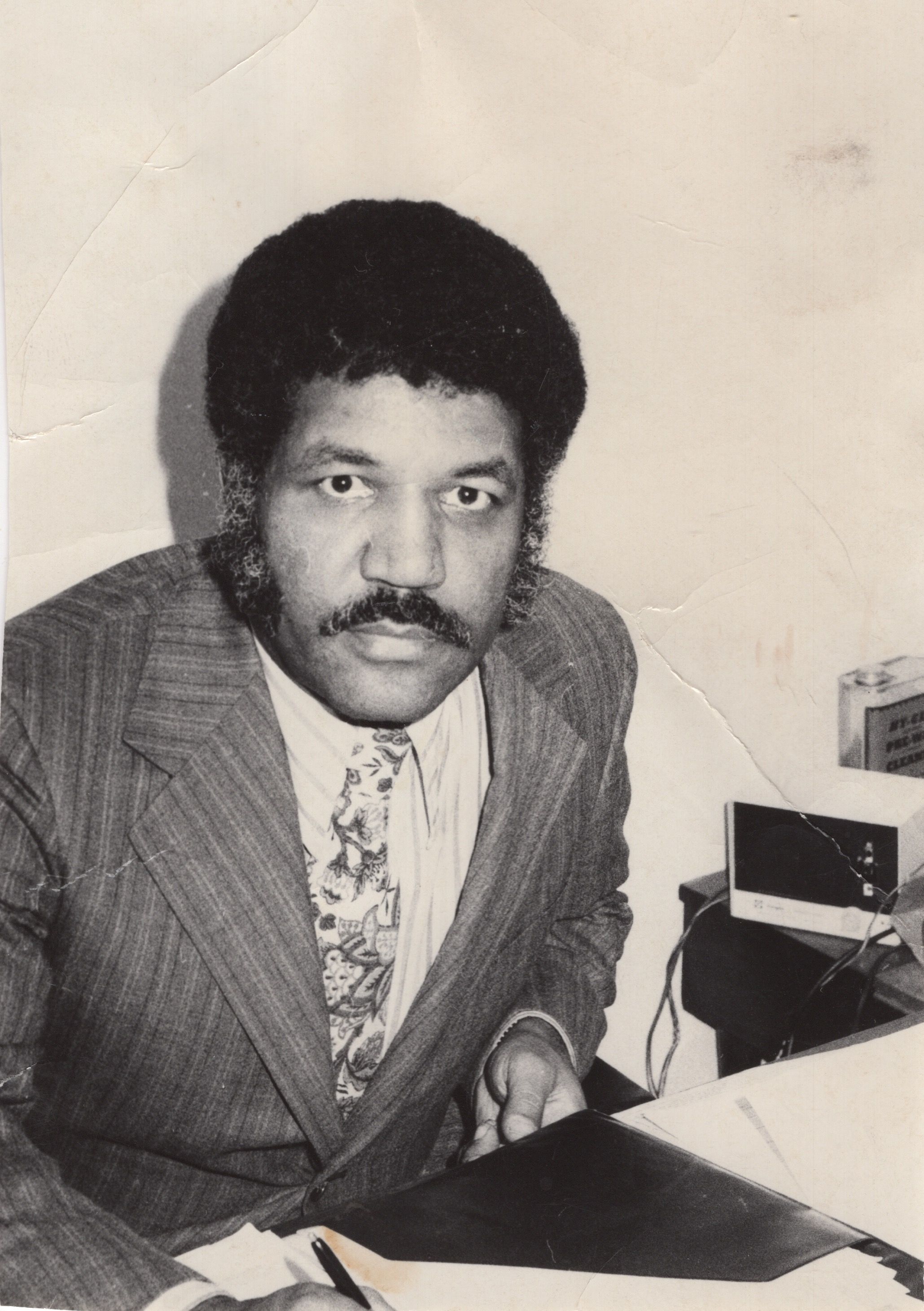 Black-and-white photo of a man with an Afro and mustache, wearing a pinstriped suit and floral tie, seated at a desk with papers and a small vintage device nearby.