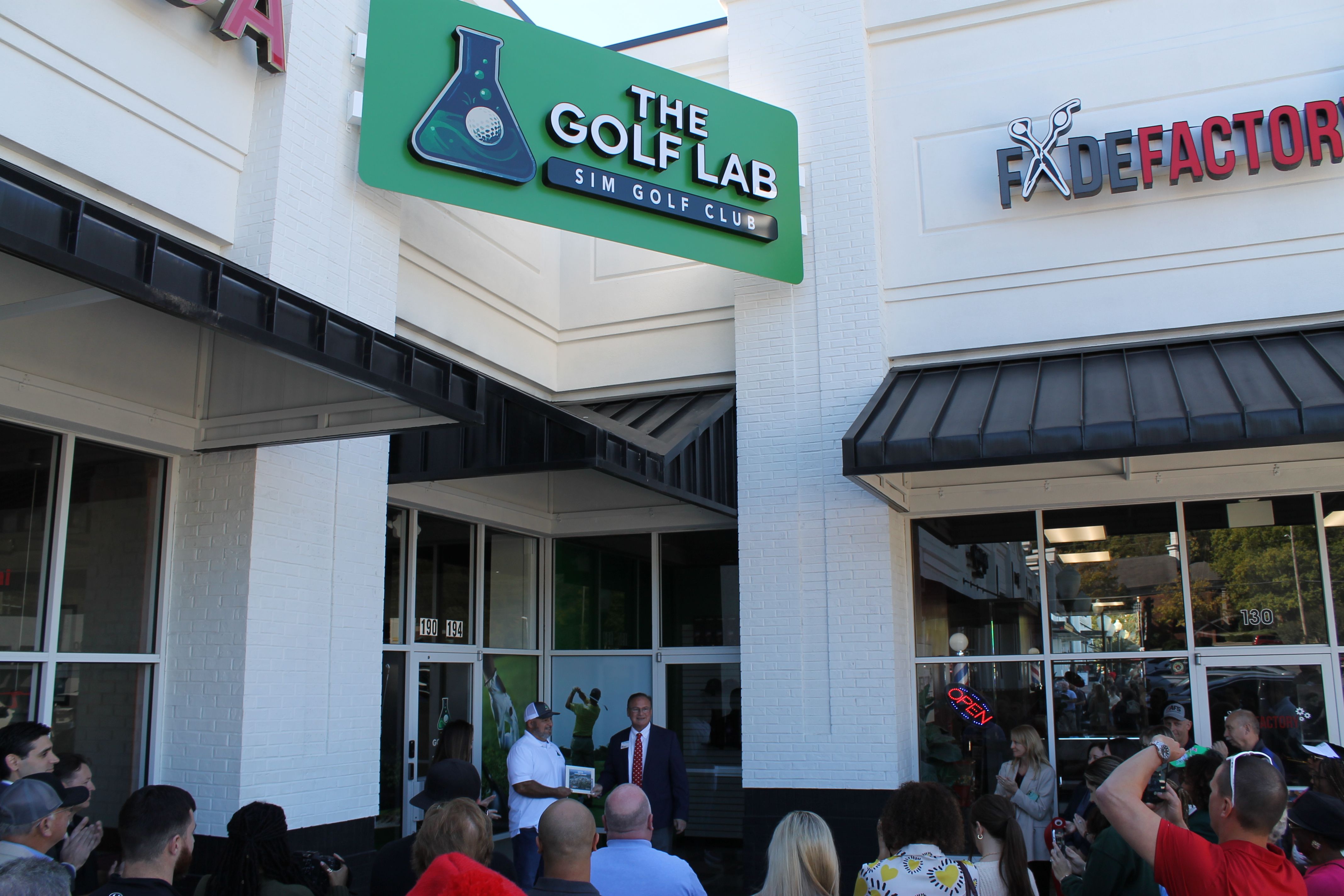 Crowd gathered outside a white brick building with black awnings for the opening of The Golf Lab Sim Golf Club, next to The Fade Factory salon with an open neon sign.