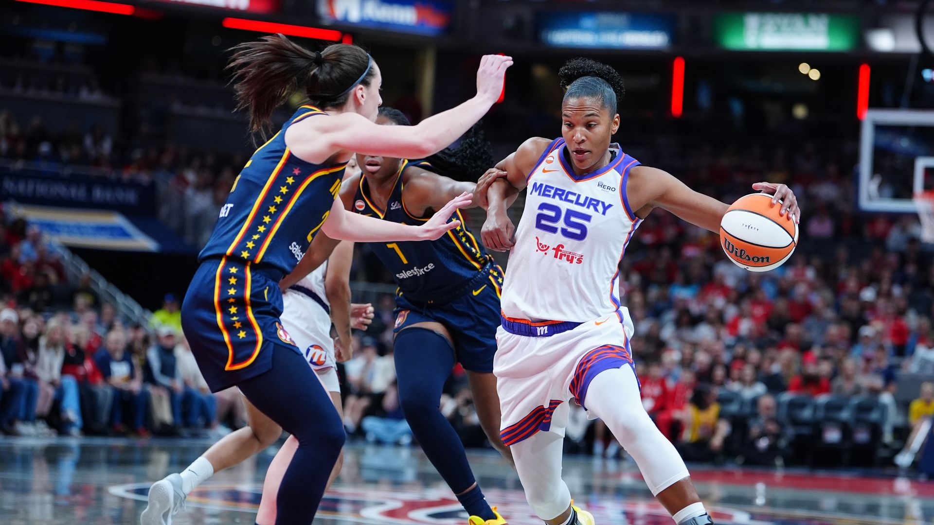 A woman in a basketball uniform with purple trim drives with the ball against a defender in a dark blue uniform with yellow trim. Players from both teams are behind them in an arena filled with seated fans. 
