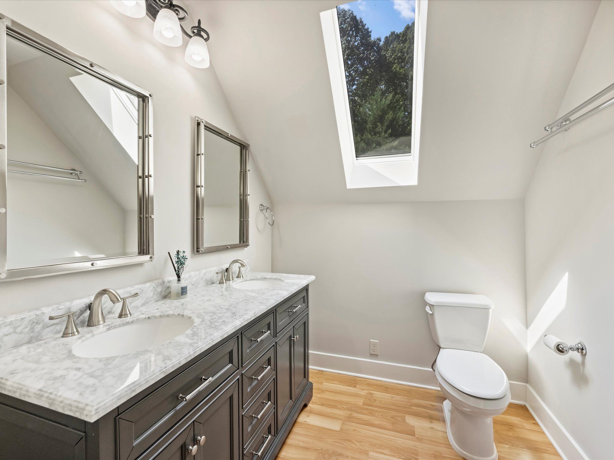 Bright bathroom with double sinks on dark wood vanity, marble countertop, two framed mirrors, skylight showing trees, white walls, hardwood floor, and a white toilet.