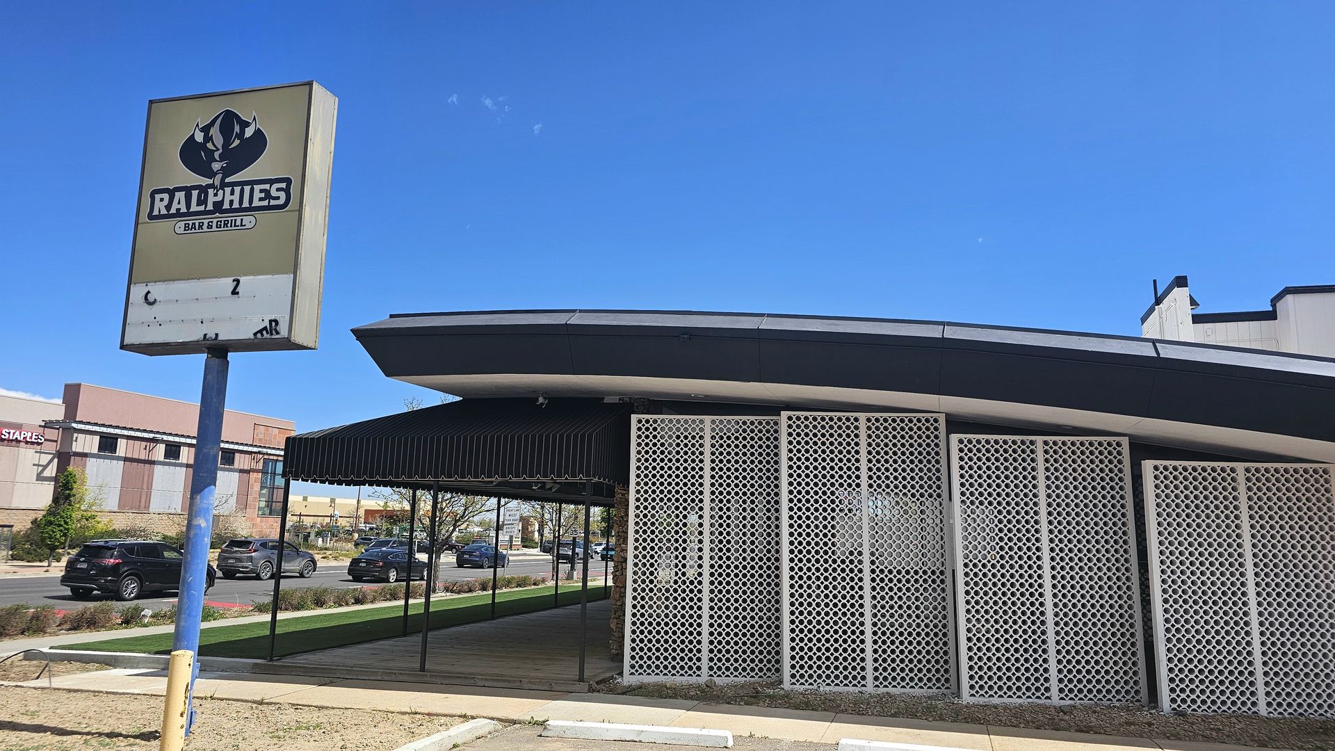 Bright day at shuttered Ralphies Bar & Grill. A tall sign on a blue pole with a horned-animal logo and the name. In front is a building with a black awning and white lattice panels; cars line the street.
