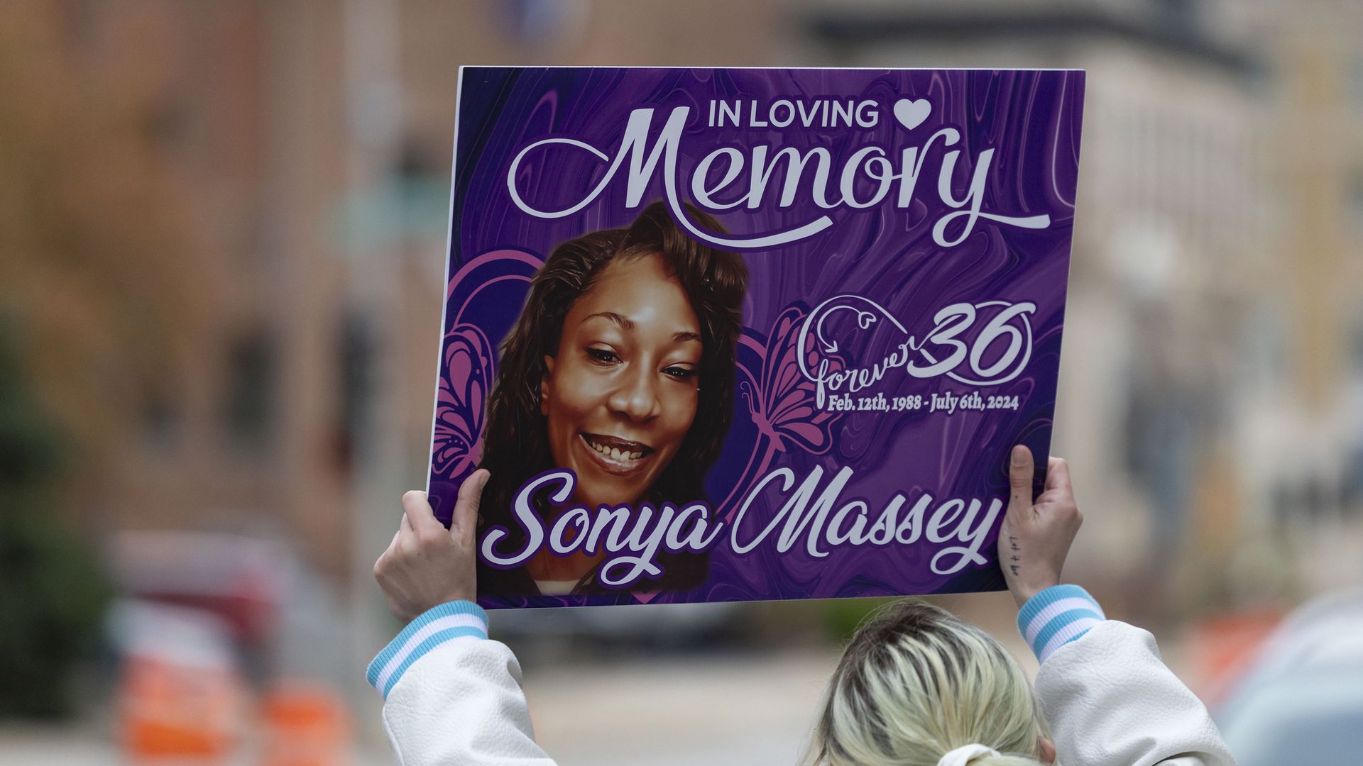 Person holding a purple memorial sign with a smiling woman's photo, text "In Loving Memory Sonya Massey," dates Feb 12, 1988 - July 6, 2024, and "forever 36" with butterfly graphics.