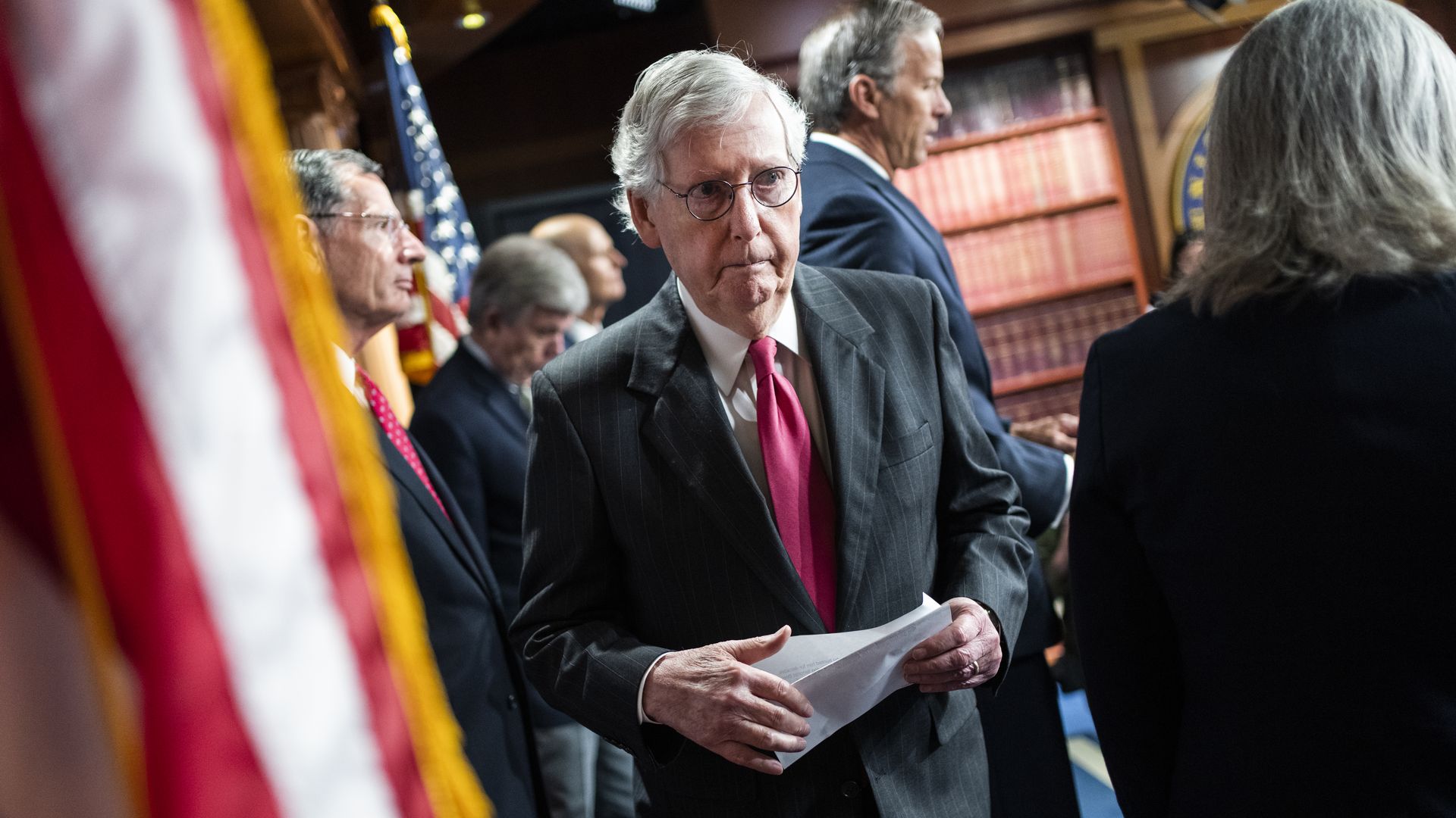 Mitch McConnell wearing a gray suit and red tie departs a press conference where multiple other Republican Senate leaders are speaking.