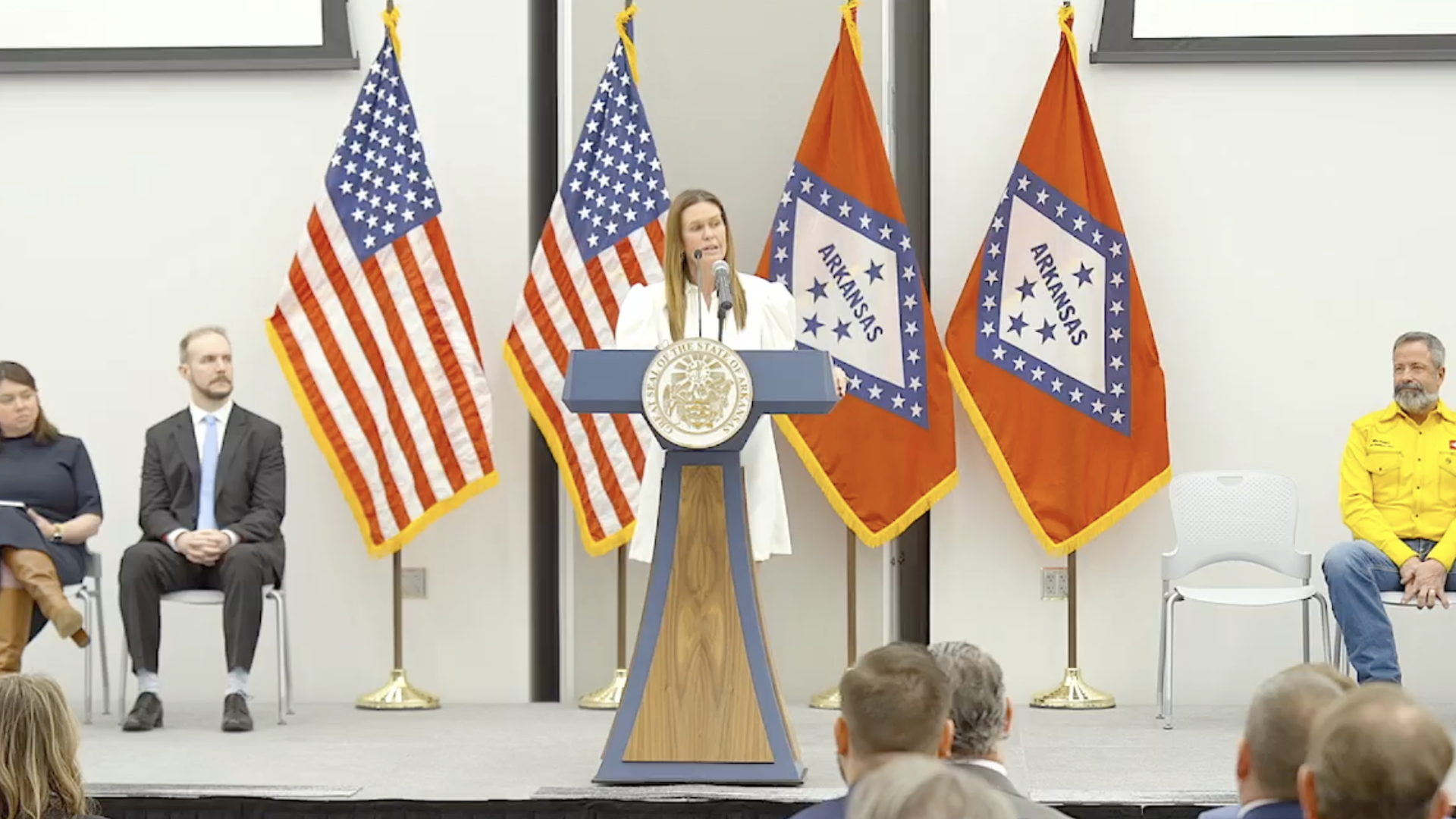 Gov. Sarah Huckabee Sanders stands behind a podium on a stage, flanked by American and Arkansas state flags. 
