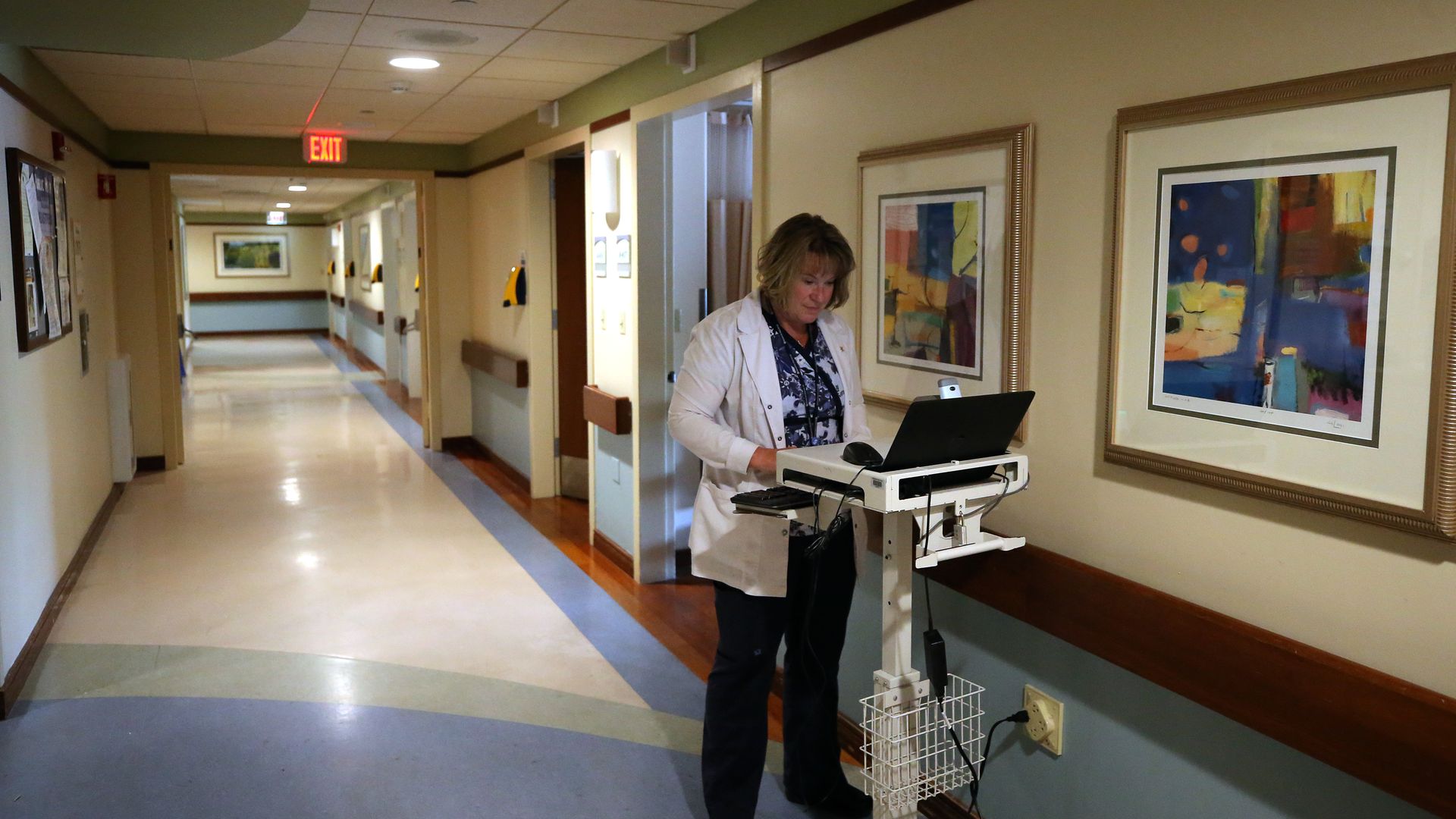A doctor works on a laptop in the hallway of a hospital.