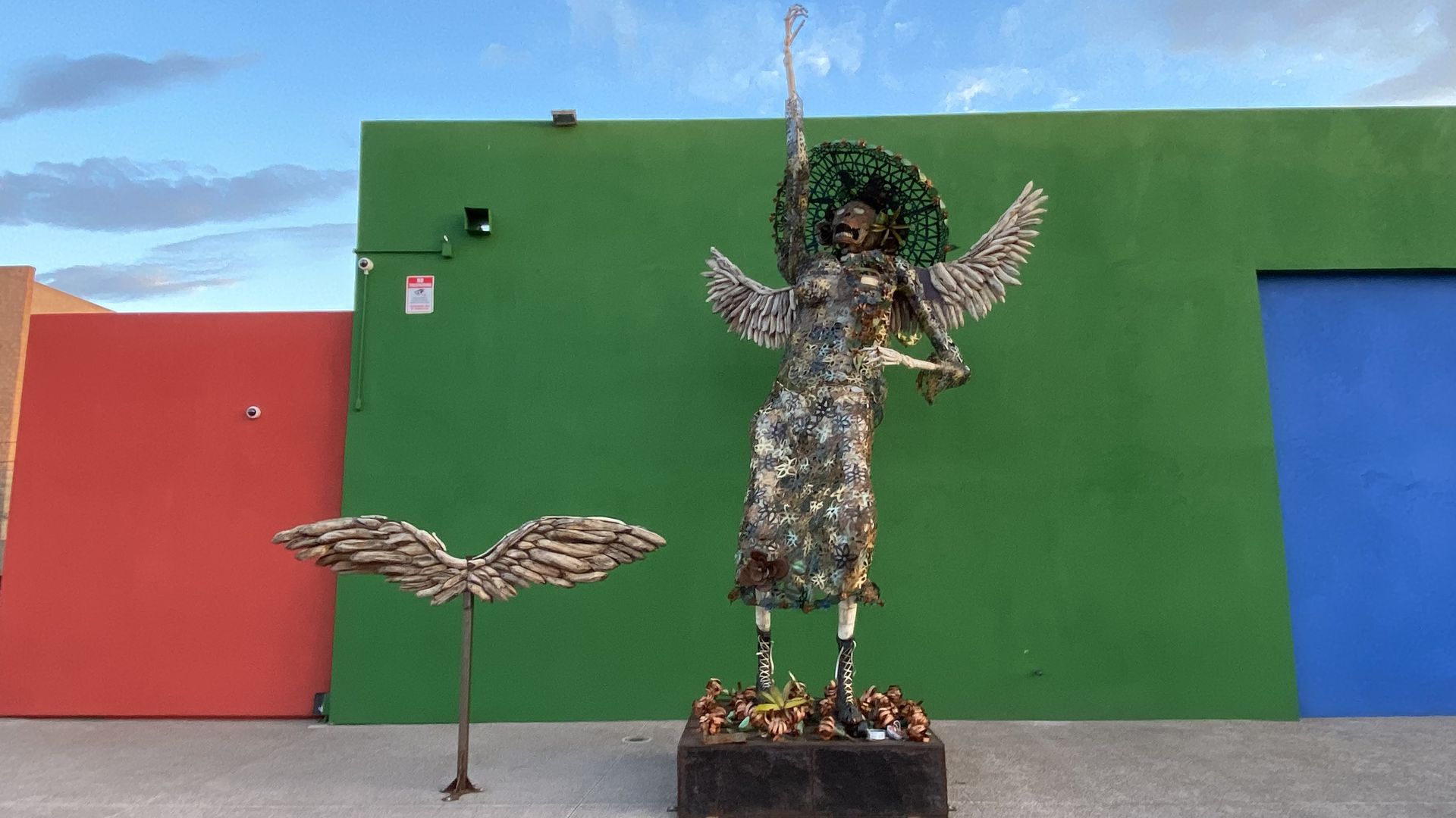 A gold colored metal statue of a skeleton in a dress with wings and a broad brimmed hat. 