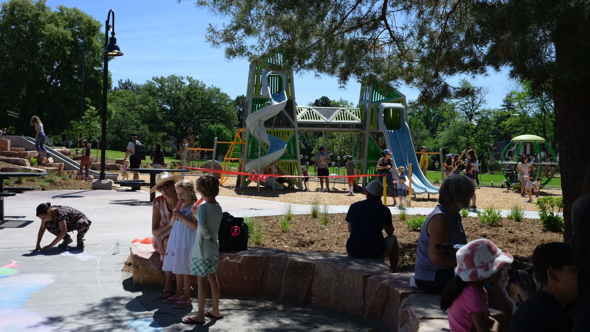 Children and adults are in the foreground in the shade near a playground, which includes two large slides. 