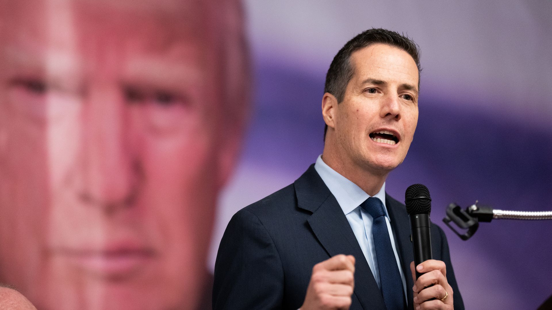 Bernie Moreno speaks into a microphone in front of a large photo of Donald Trump. 