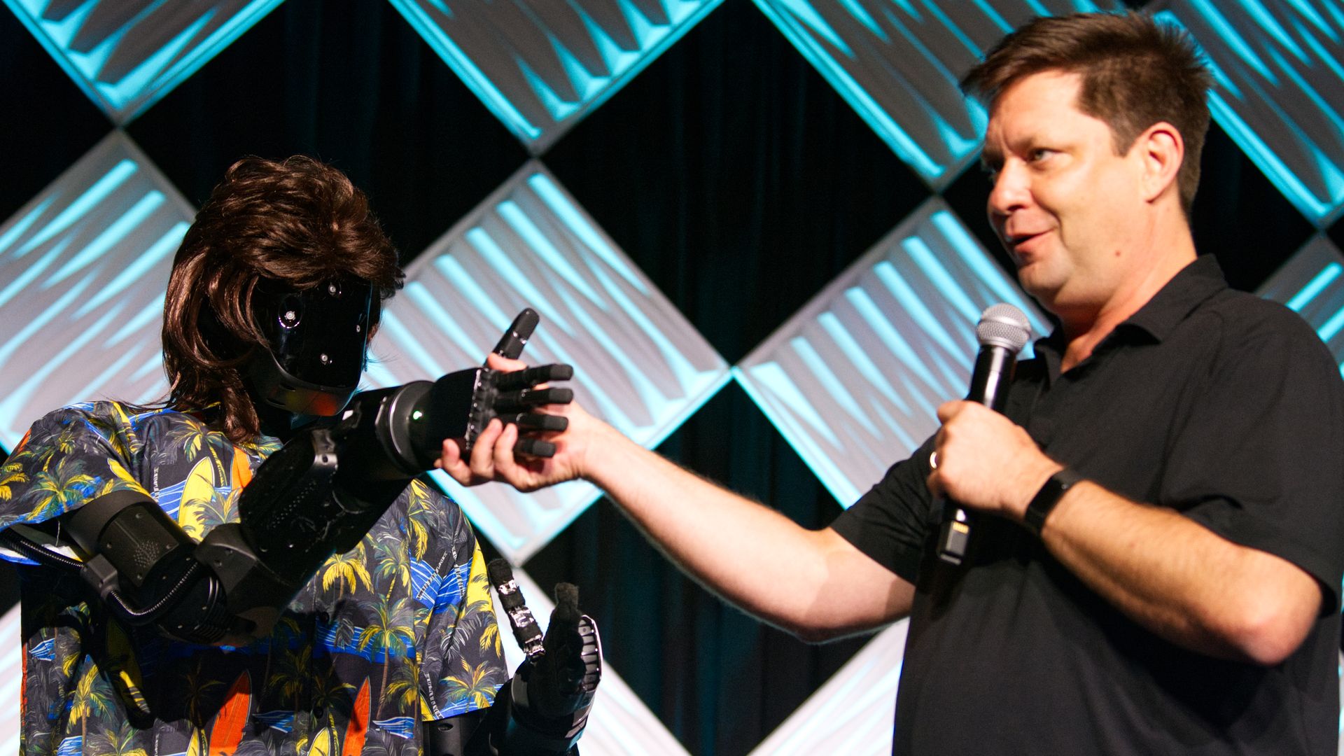A humanoid-looking robot wearing a mullet wig shakes hands with a man holding a mic on a stage.