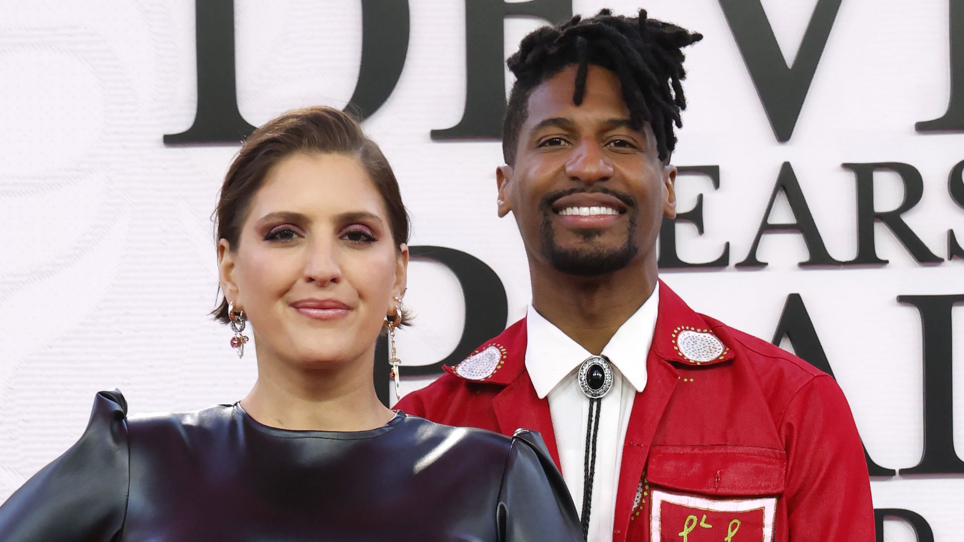 Suleika Jaouad and Jon Batiste smile on the red carpet. A sign behind them says "Devils Wears Prada."