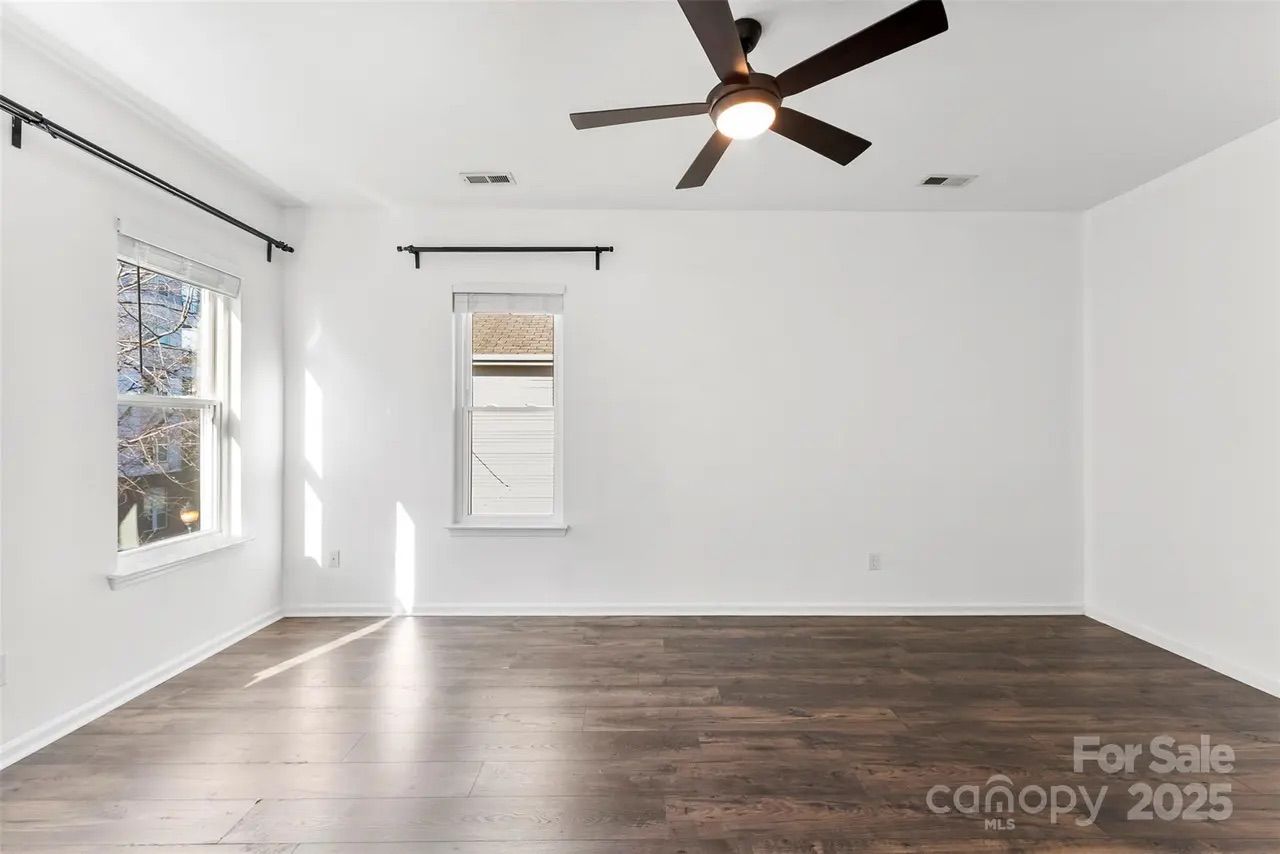 Empty room with white walls, two windows with black curtain rods, dark wood flooring, and a black ceiling fan with light turned on.