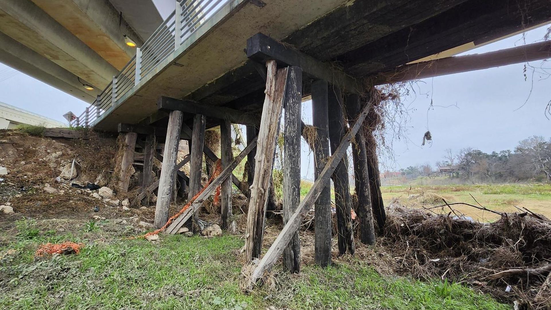 Underside of a wooden bridge supported by damaged wooden beams with greenery and dry vegetation on the ground under overcast sky.
