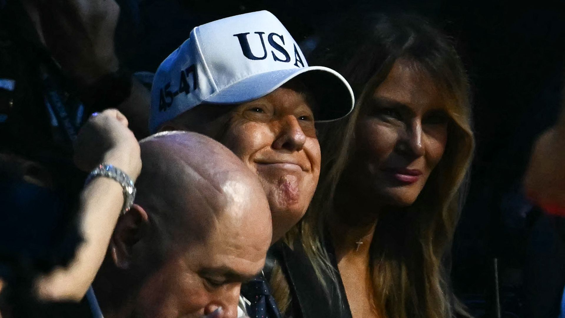 President Trump wears a white hat with the text "USA" on it while he smiles and sits next to Melania Trump.