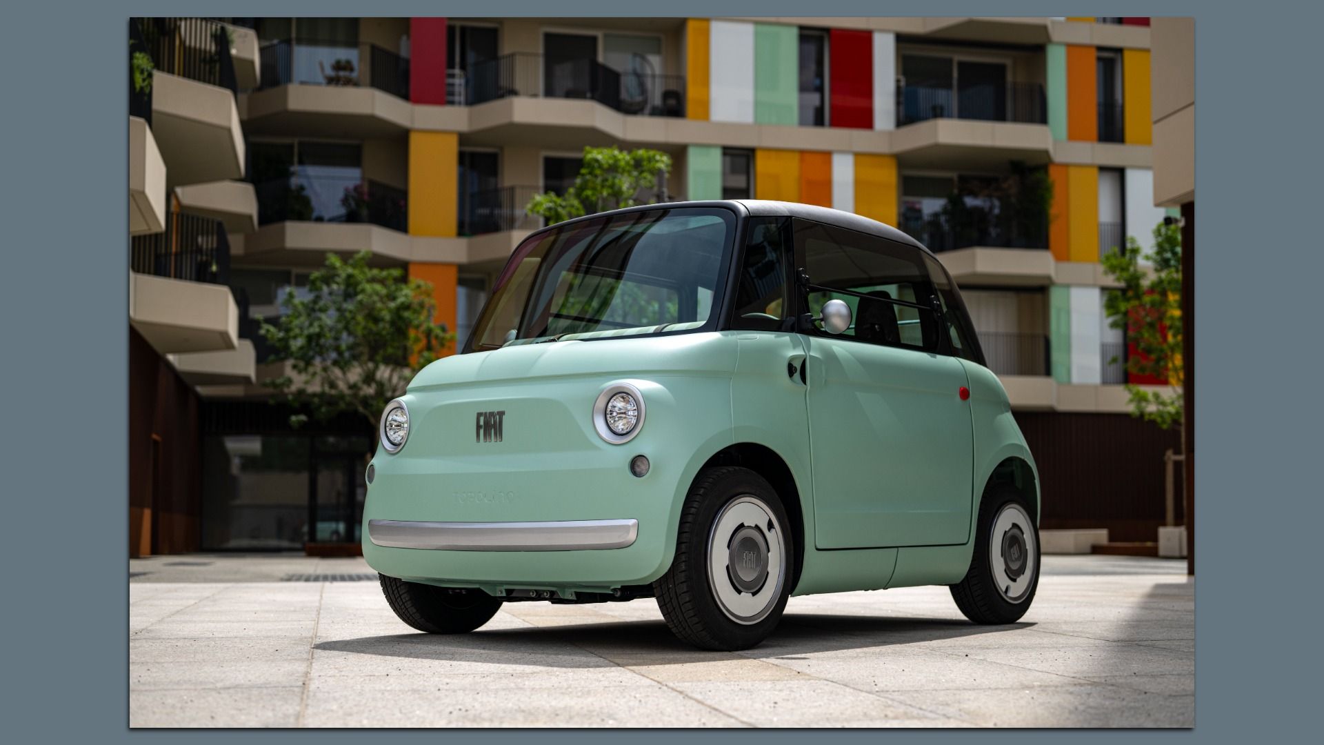 Light green Fiat Topolino micro car with black roof and round headlights parked on pavement in front of a colorful building. 