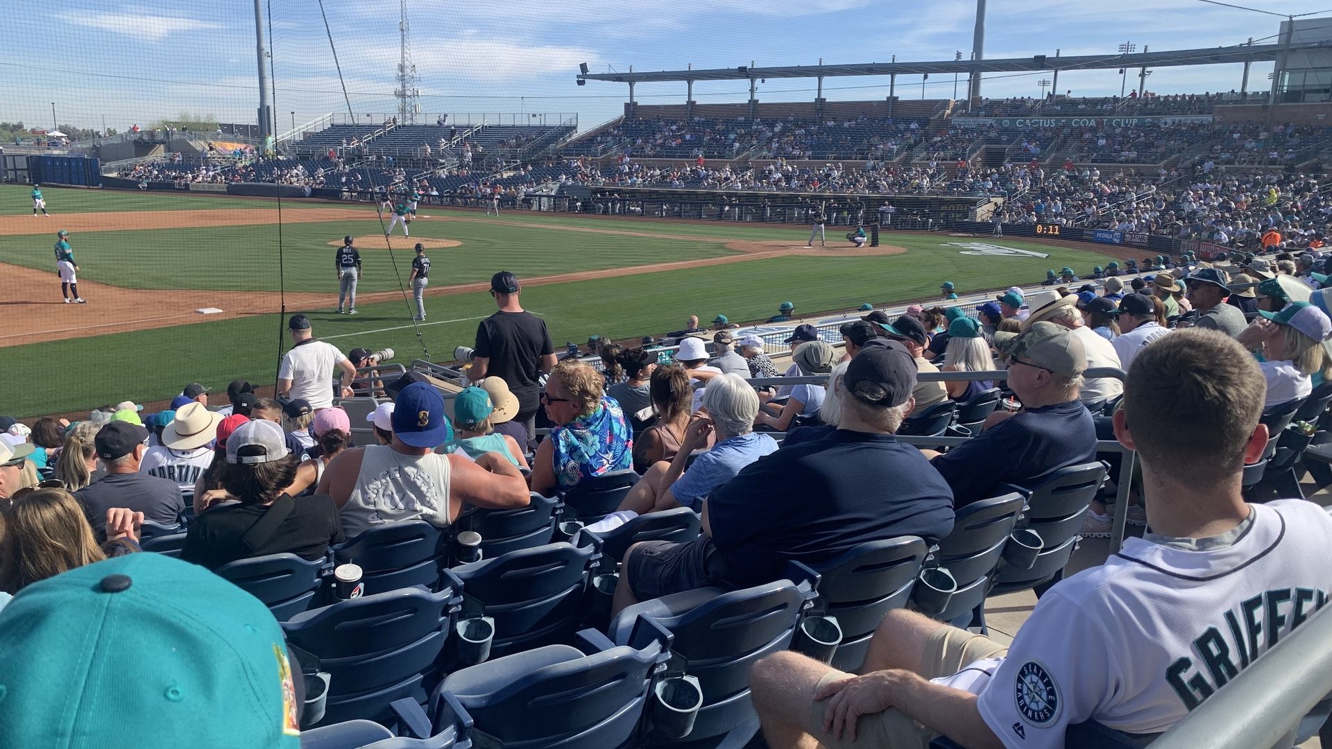 Crowded baseball stadium with fans watching a game on a sunny day, with one fan wearing a white Seattle Mariners jersey labeled Griffey.