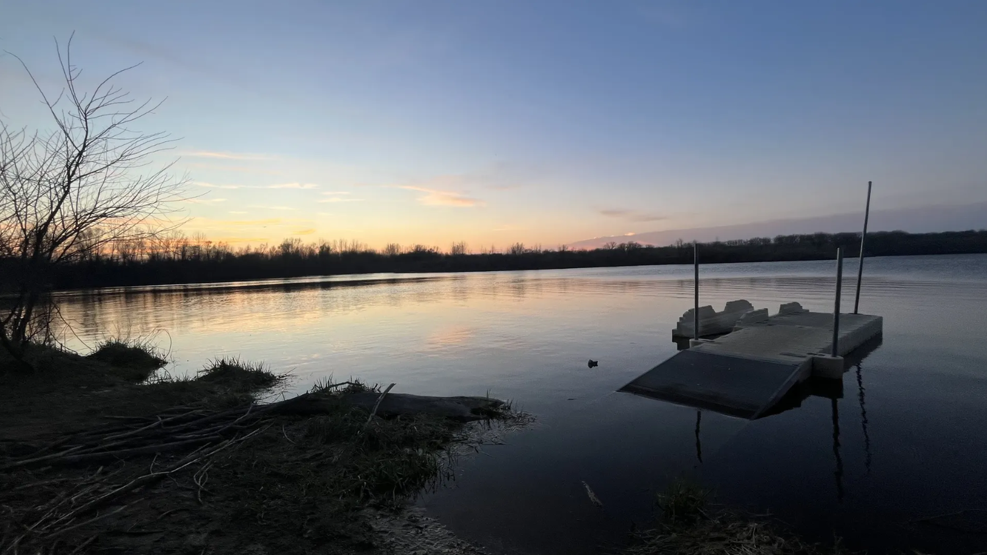 Calm lake at sunset with a floating dock on the right, reflecting orange and blue skies; bare trees line the distant shore, while driftwood and branches lie along the foreground.
