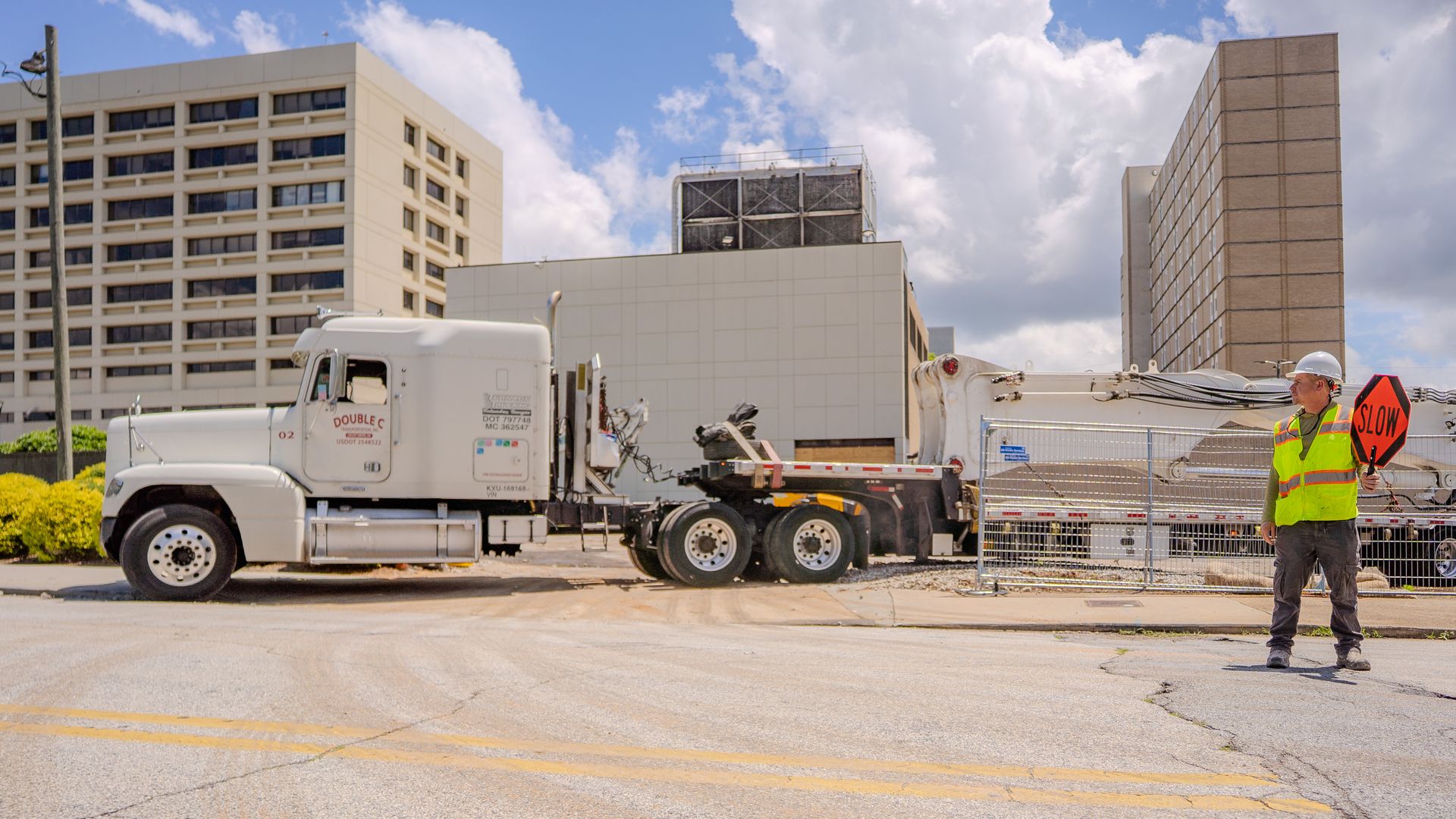An individual wearing a construction safety vest holds a "SLOW" sign as a semi truck with a long flatbed trailer maneuvers into a work site