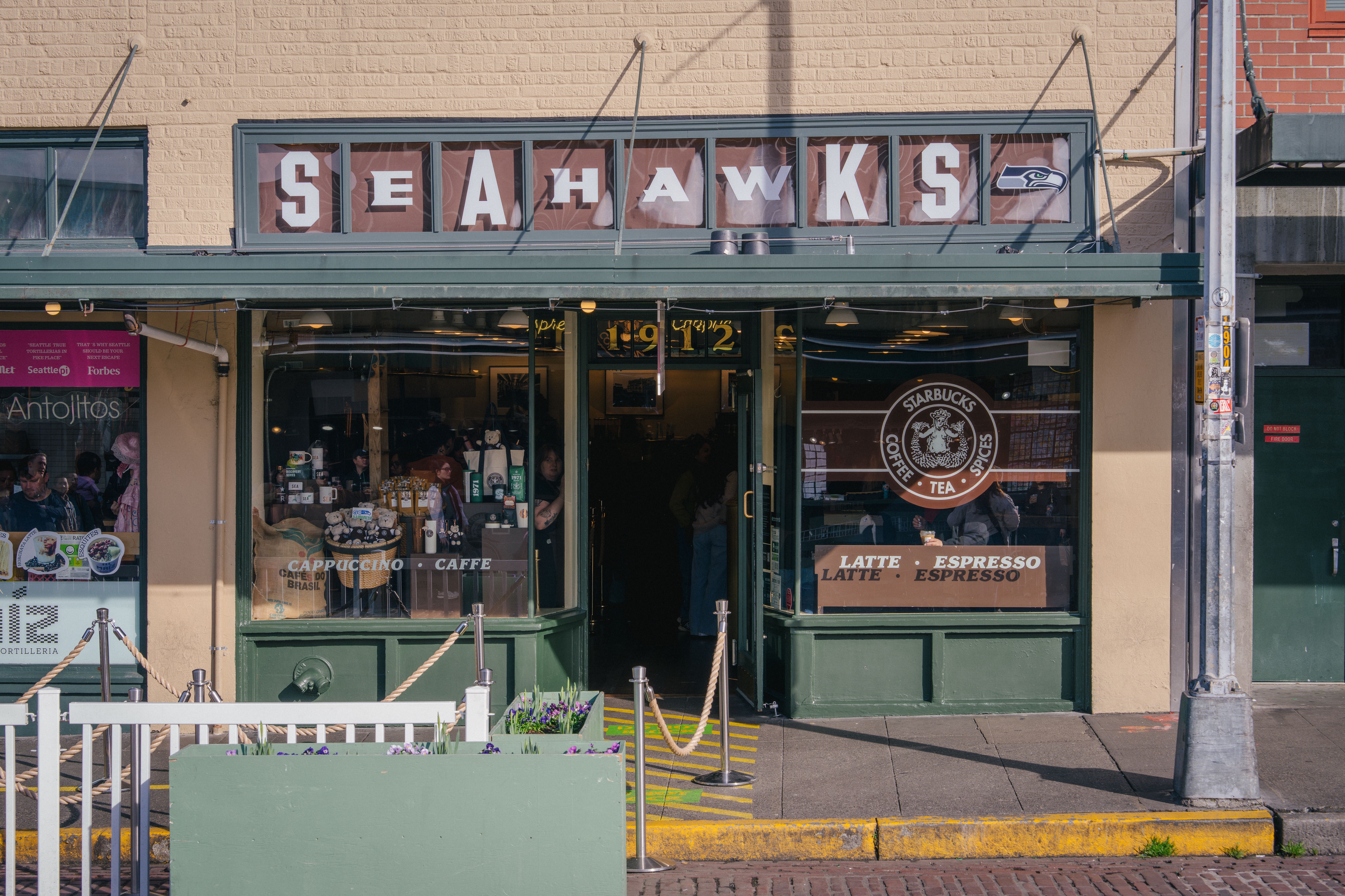 Exterior of a coffee shop with green trim and a sign reading "SEAHAWKS" above. Windows display Starbucks logo and coffee options like cappuccino, latte, and espresso. Yellow curb outside.