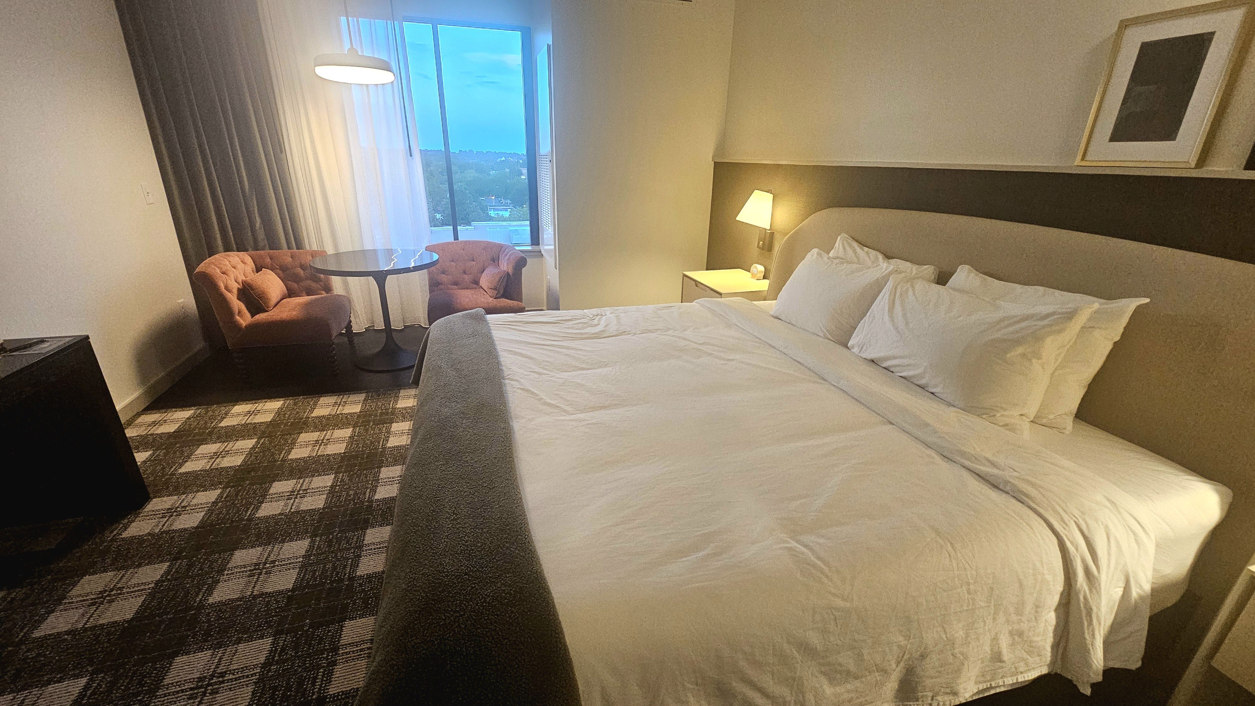 Hotel room with a large white bed on the right, pillows and a bedside lamp. A window nook features two pink tufted chairs around a round table, with a blue evening view outside.