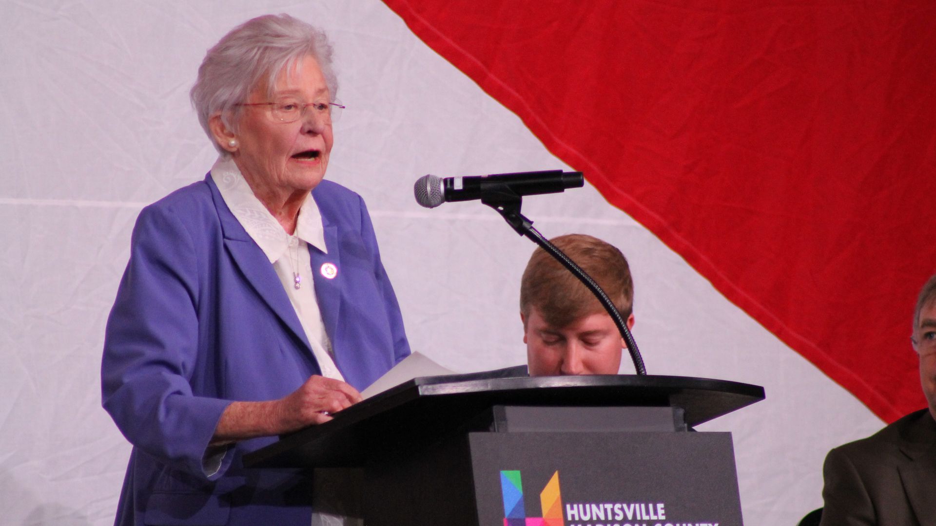 An elderly woman with short white hair and glasses, in a purple blazer, speaks into a microphone at a podium against a red and white backdrop, with a Huntsville Madison County Chamber sign on the podium.