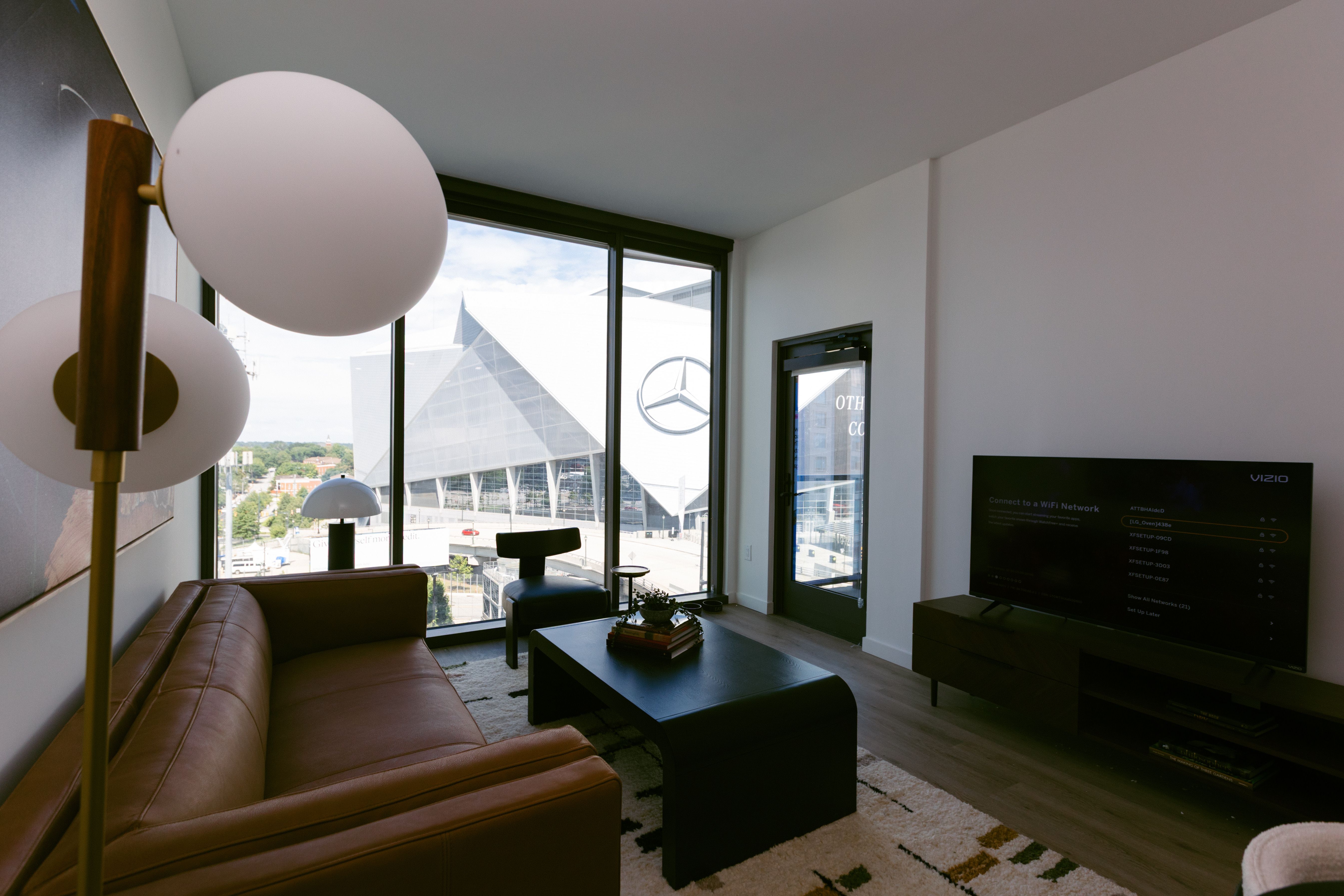 Modern living room with brown leather sofa, black coffee table with books and plant, floor lamp, and large windows showing a view of Mercedes-Benz Stadium.
