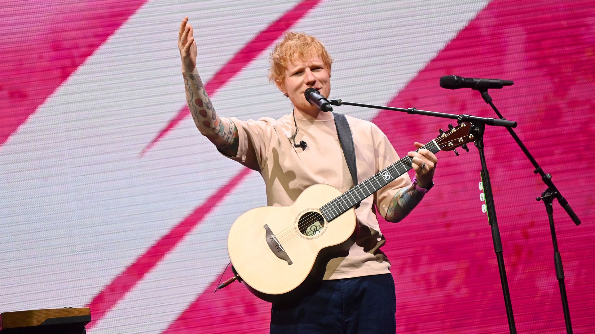 Male singer with blond hair and tattooed arms plays an acoustic guitar on stage, singing into a microphone with one hand raised, a pink diagonal backdrop behind him.