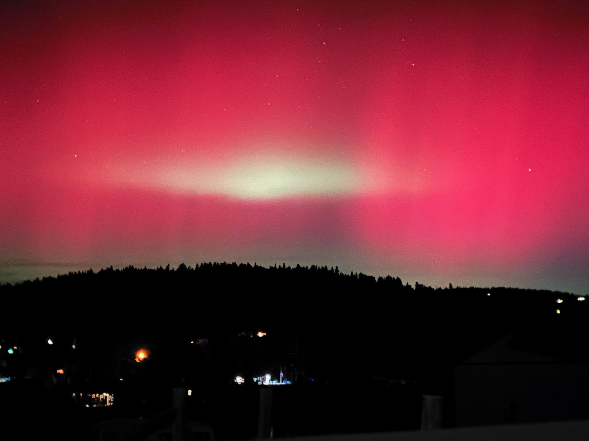 Night sky with vibrant red and white northern lights above a dark forest silhouette and scattered city lights below.