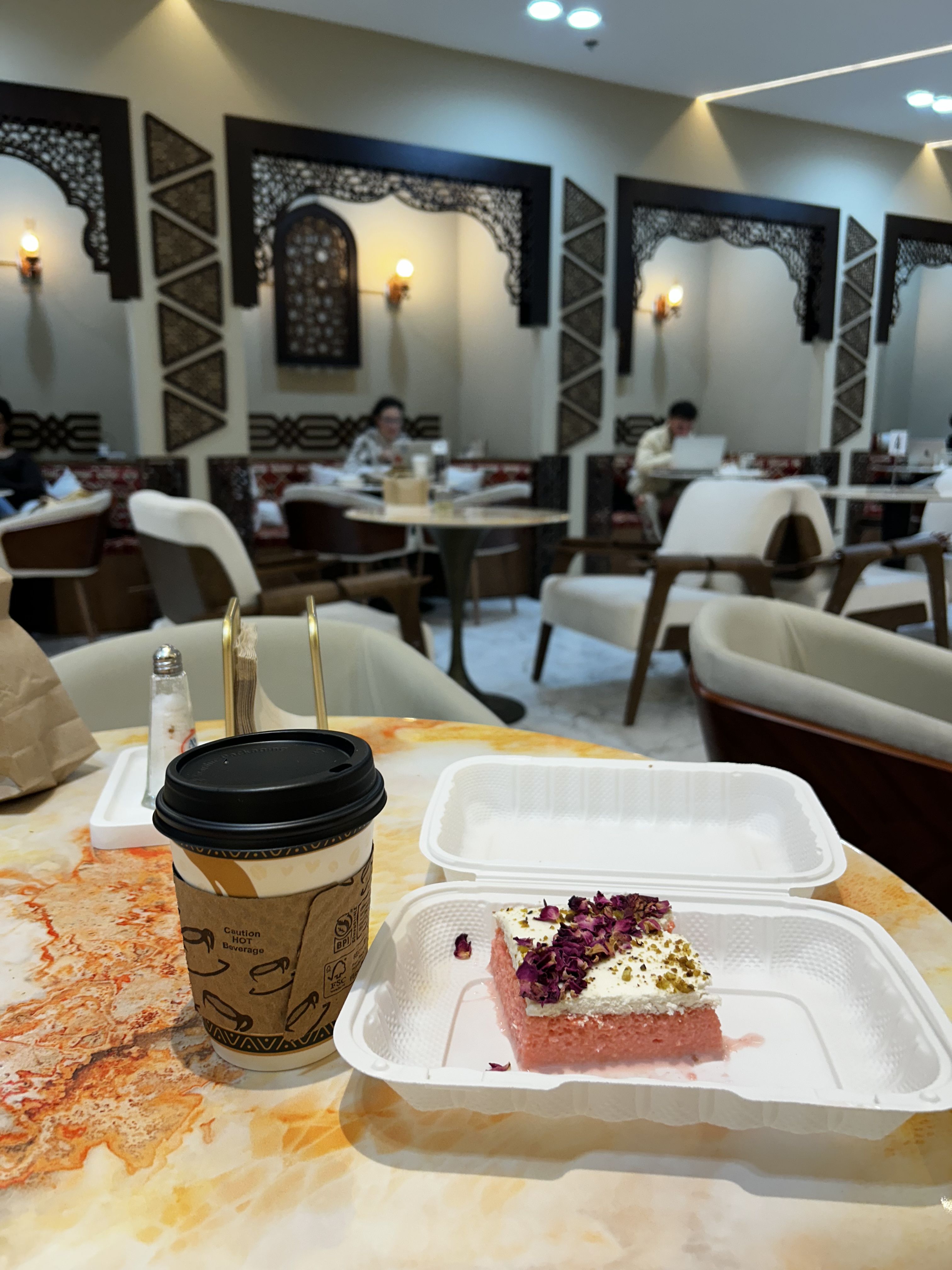 Table with a takeaway cup of coffee and a pink dessert topped with white cream and rose petals in a white container, inside a café with ornate wooden wall decorations and soft lighting.