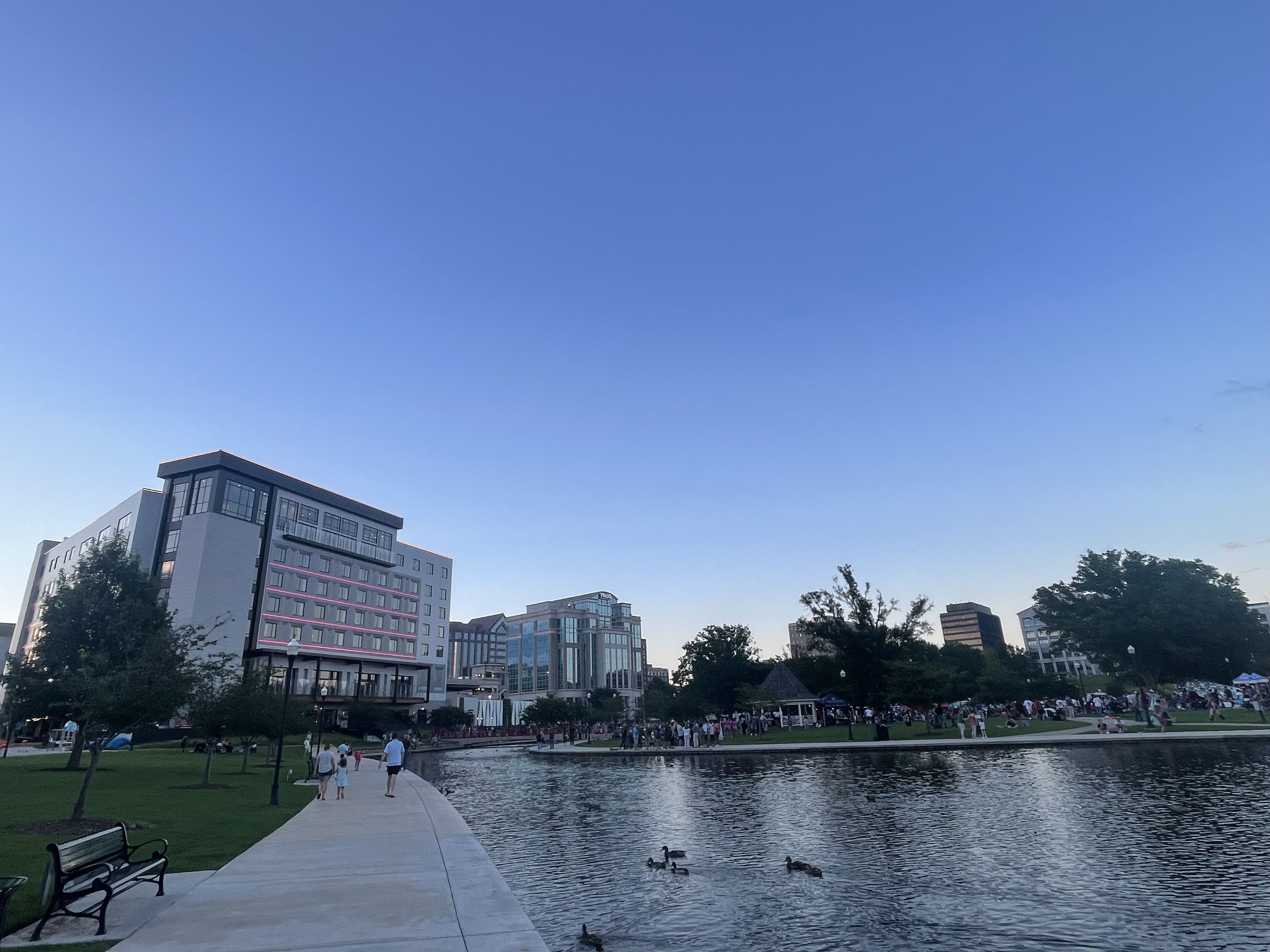 Urban park at dusk with people walking on path beside lake with ducks, modern buildings on left, and crowd gathered near trees and gazebo under clear blue sky.