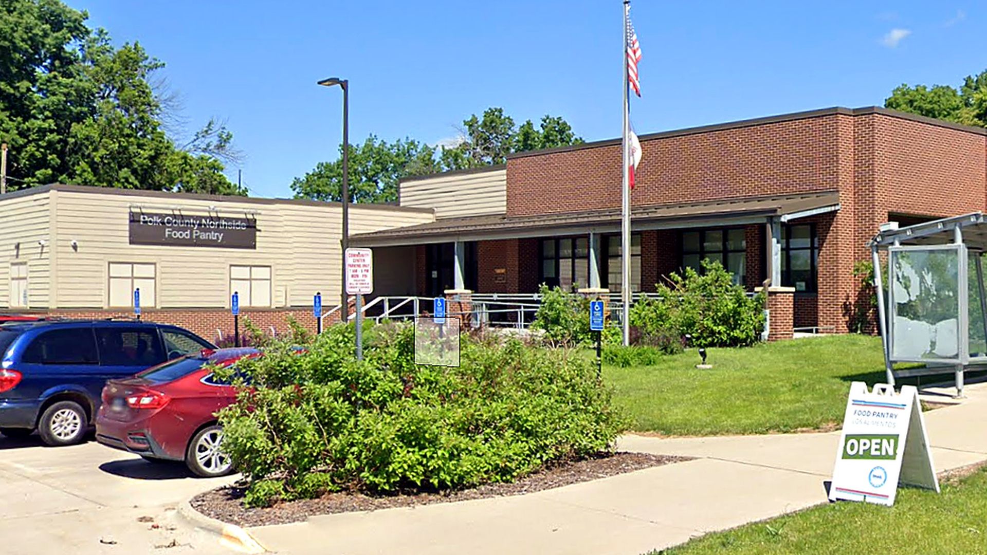 Sunlit, single-story building labeled "Food Pantry" with a flagpole, ramp, handicapped parking, shrubs, and several cars; an OPEN sign on the sidewalk.