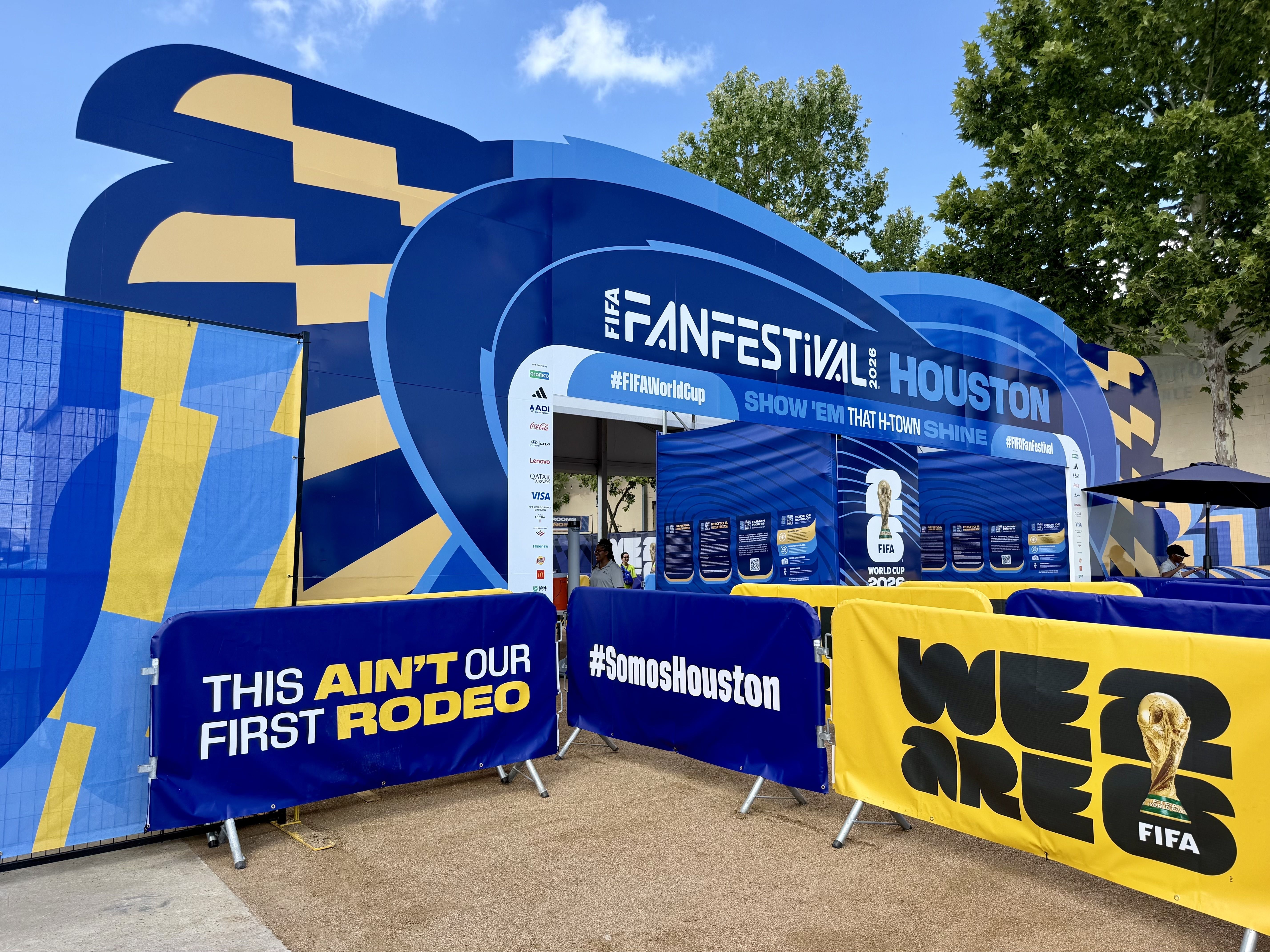 FIFA Fan Festival in Houston with blue and yellow arches, large banners, and crowd barriers along a walkway; clear blue sky and green trees in the background.