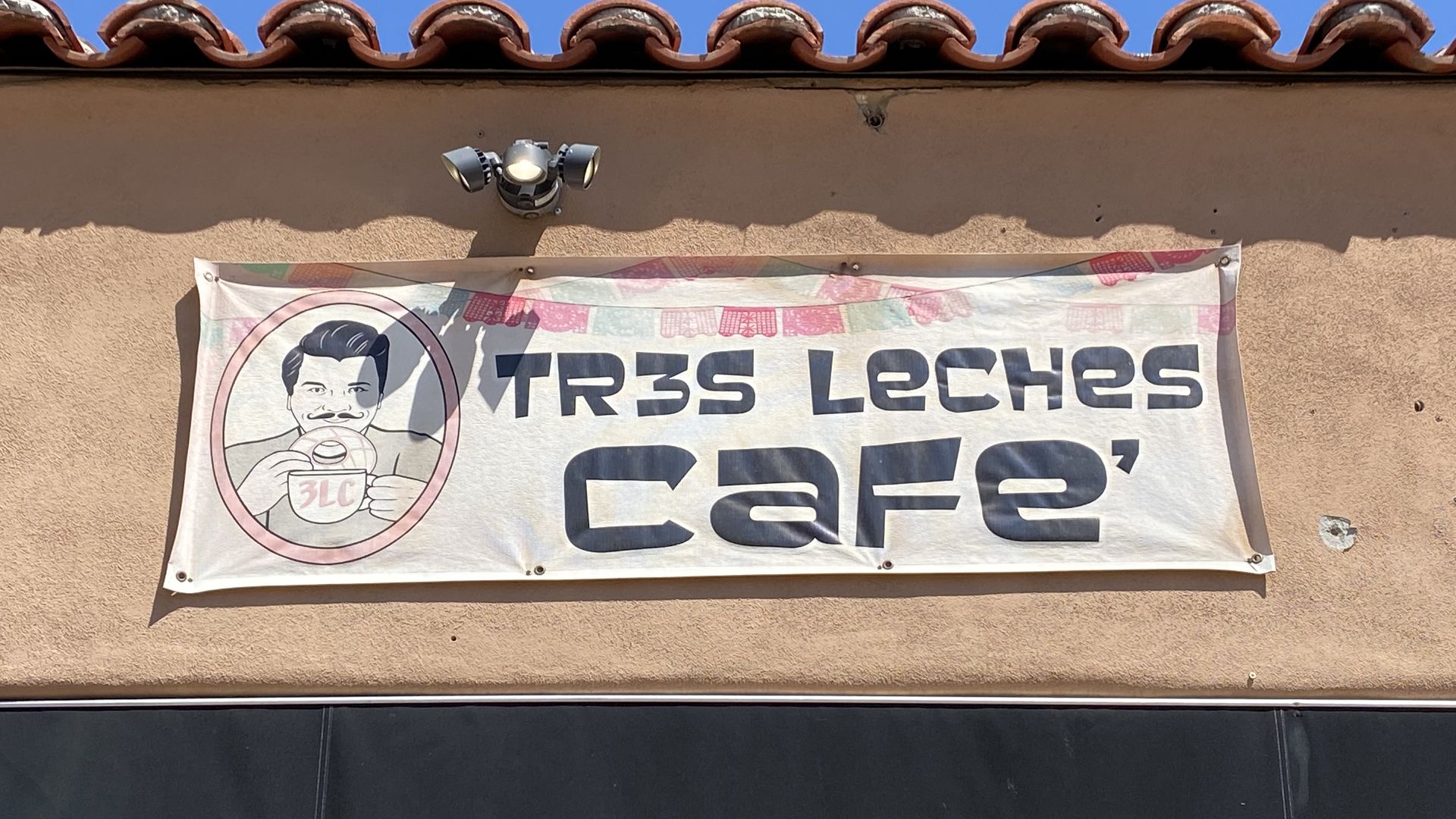Bright blue sky above a beige stucco wall with curved terracotta roof tiles. A silver outdoor light fixture sits over a banner showing a cartoon man holding a cup and the words "Tres Leches Café".