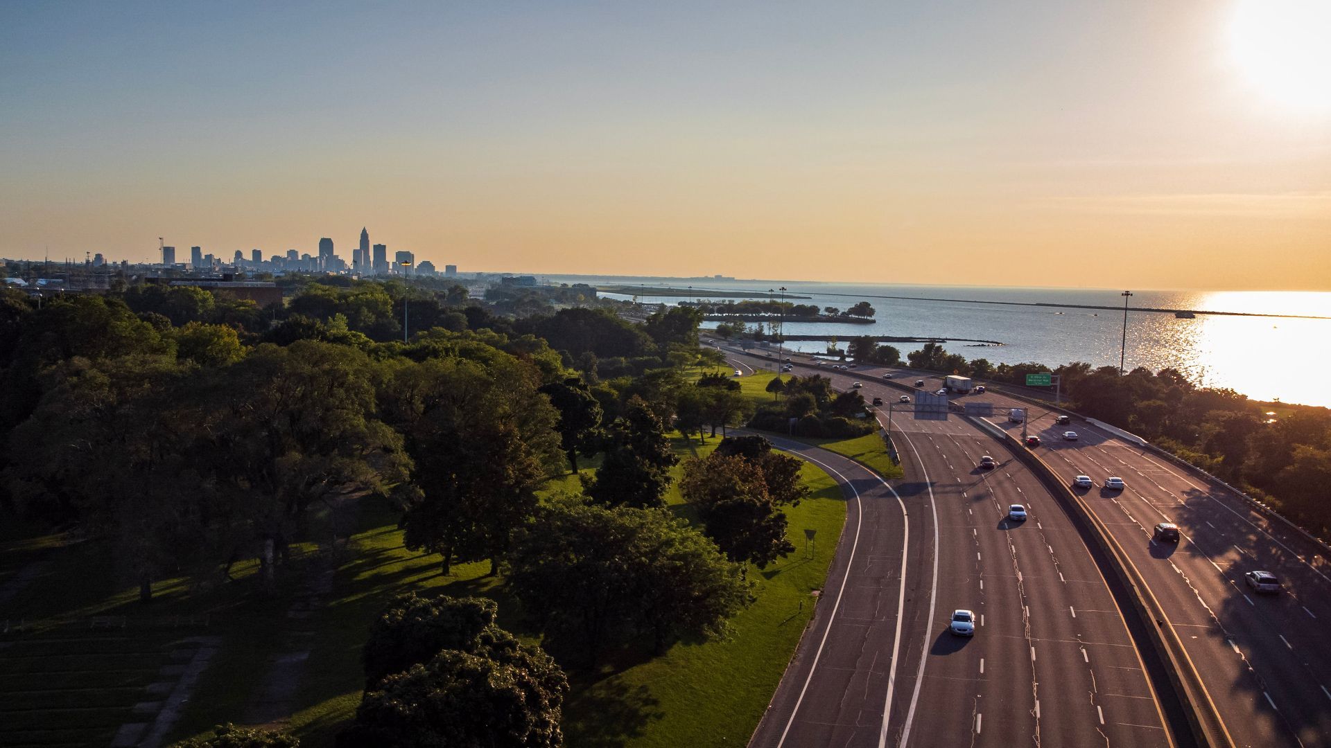 Aerial view of a coastal city at sunset: skyline along the left horizon, calm water to the right, a winding highway with cars, and lush green trees and a park in the foreground.