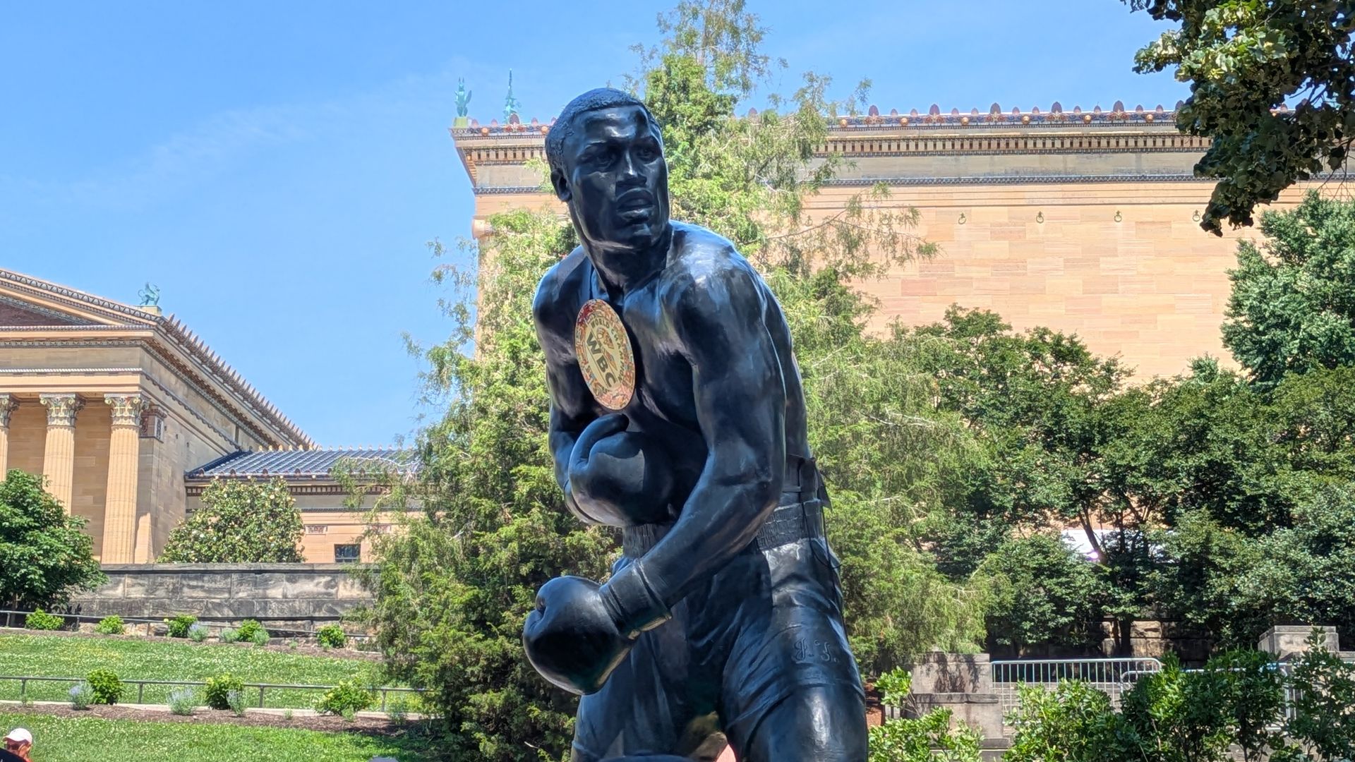 Bronze statue of a man in a dynamic running pose with a round emblem on his chest, in sunny park. Behind: classical buildings with columns, trees, and a clear blue sky; several people sit nearby.