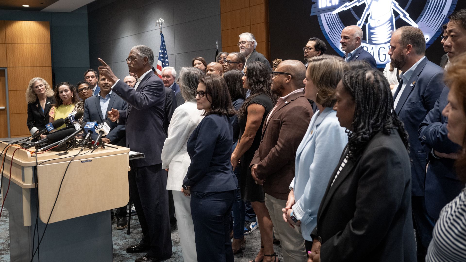 A diverse group of people stand attentively in a conference room; a man in a suit speaks at a podium with many microphones. An American flag and an IBEW logo are visible in the background.