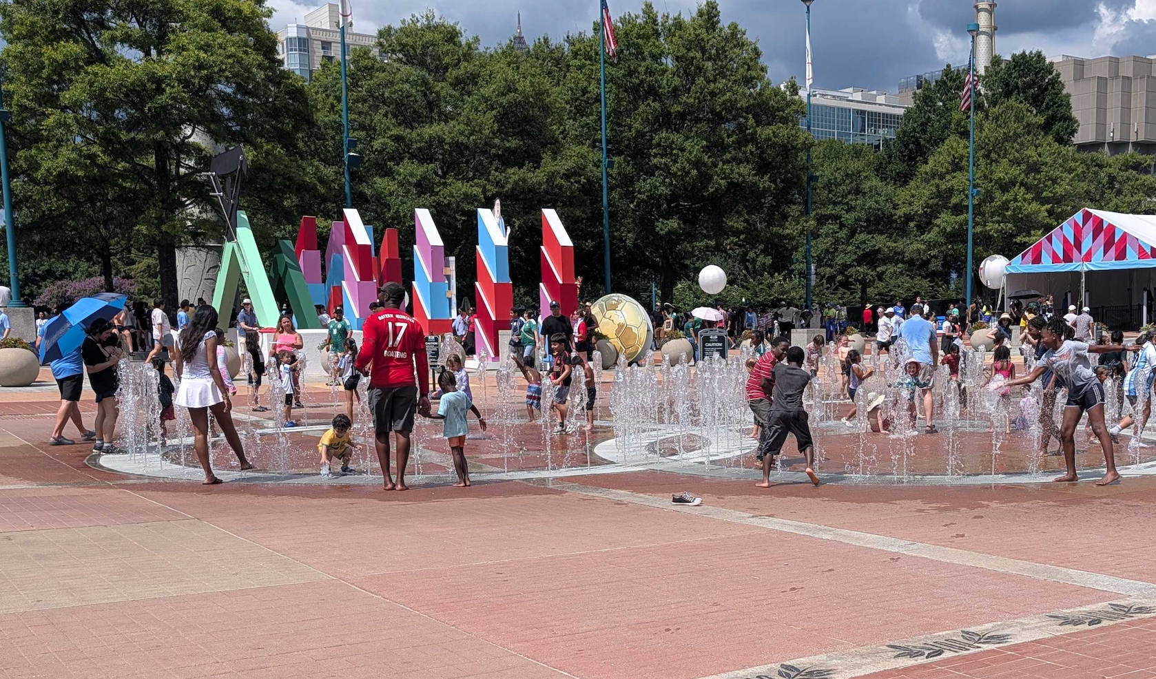 Children and adults splash in a circular fountain in a sunny plaza; vibrant geometric sculptures rise behind them, with trees, flags, and a striped tent on the right.