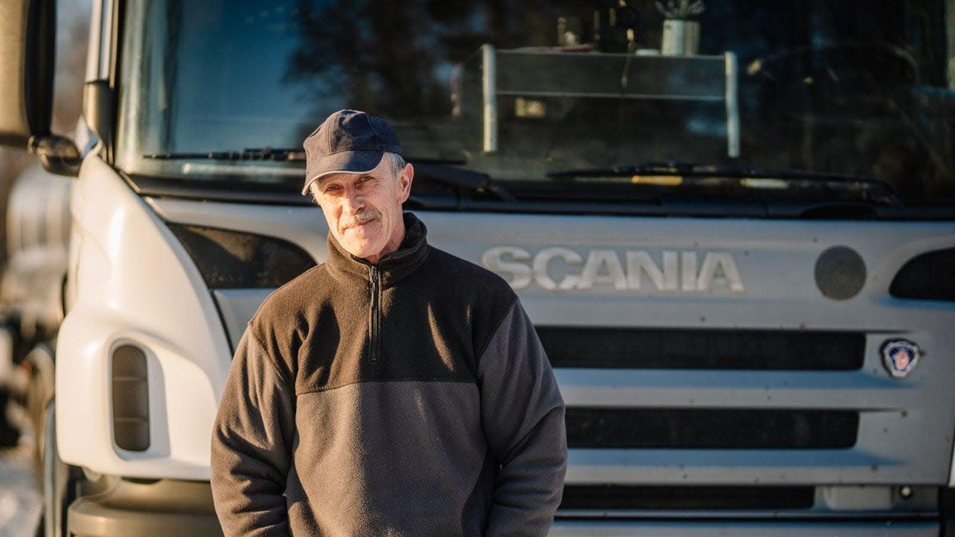 Middle-aged man wearing a black cap and two-tone fleece jacket standing in front of a white Scania truck during daylight.