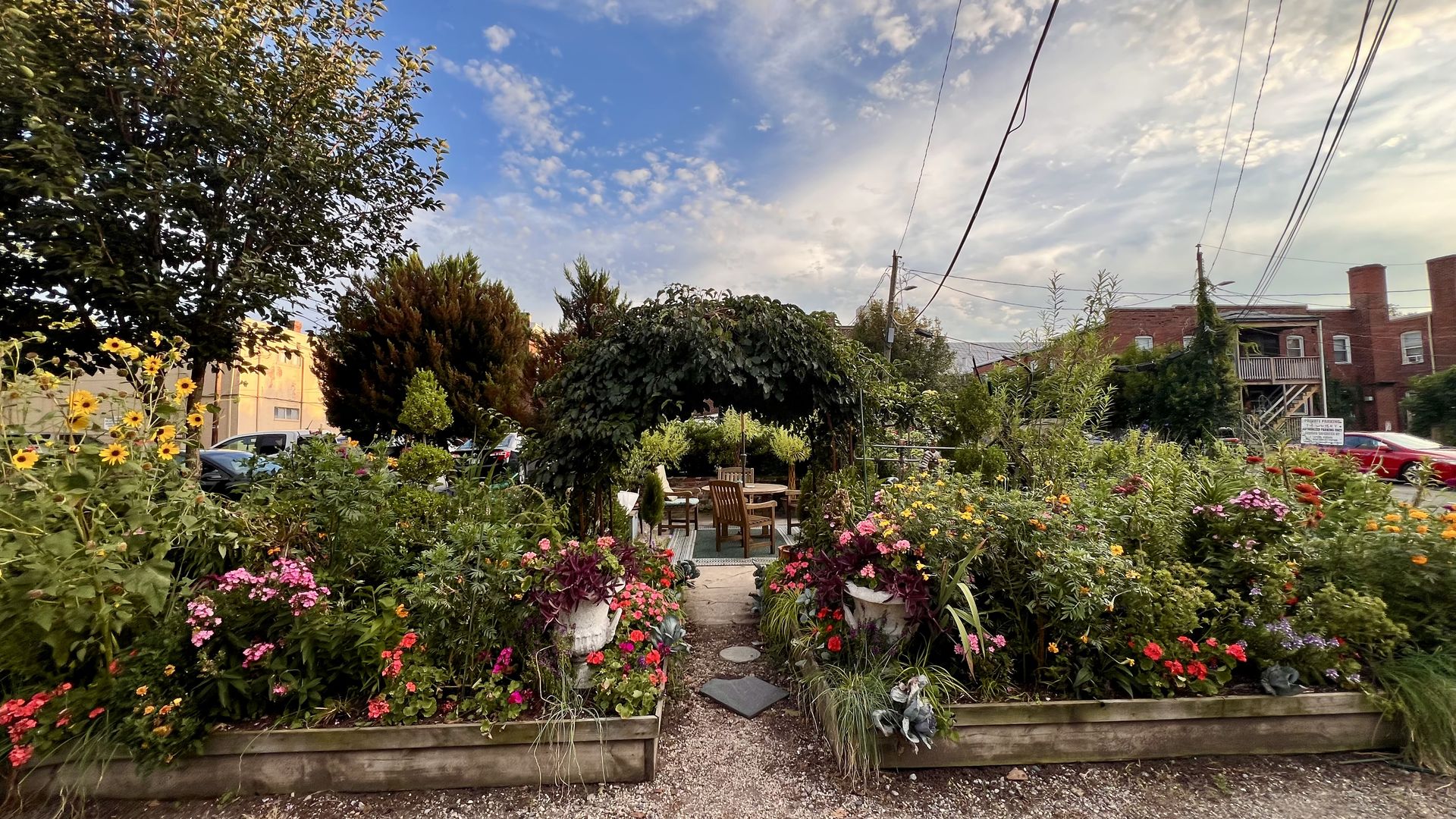 Vibrant community garden with a variety of colorful flowers and plants, two large white planters, a leafy archway, wooden chairs and table, set against a partly cloudy blue sky.