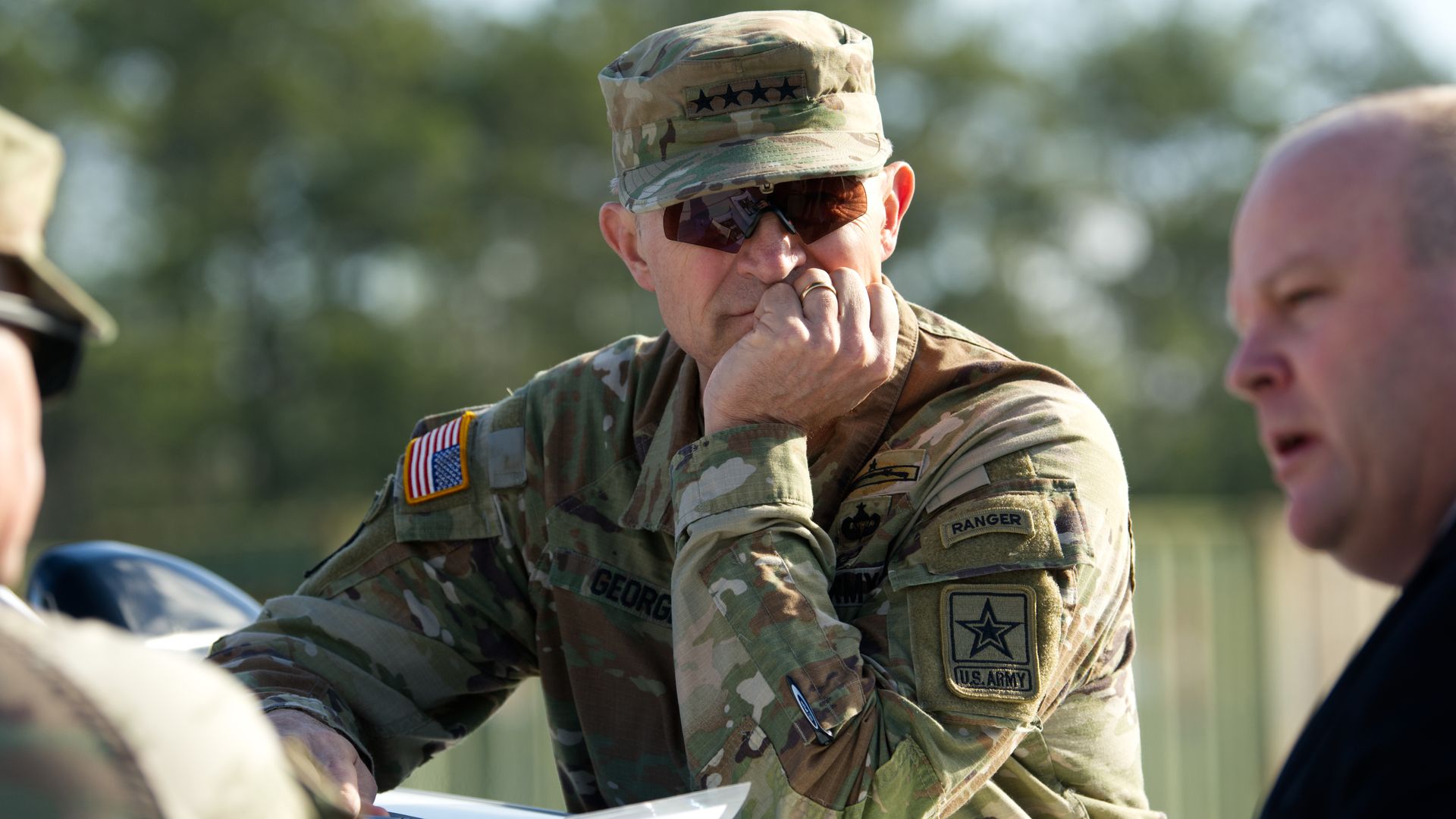 An Army officer in camouflage uniform and cap with stars, wearing dark sunglasses, rests his chin on his hand while looking at papers on a table outdoors.