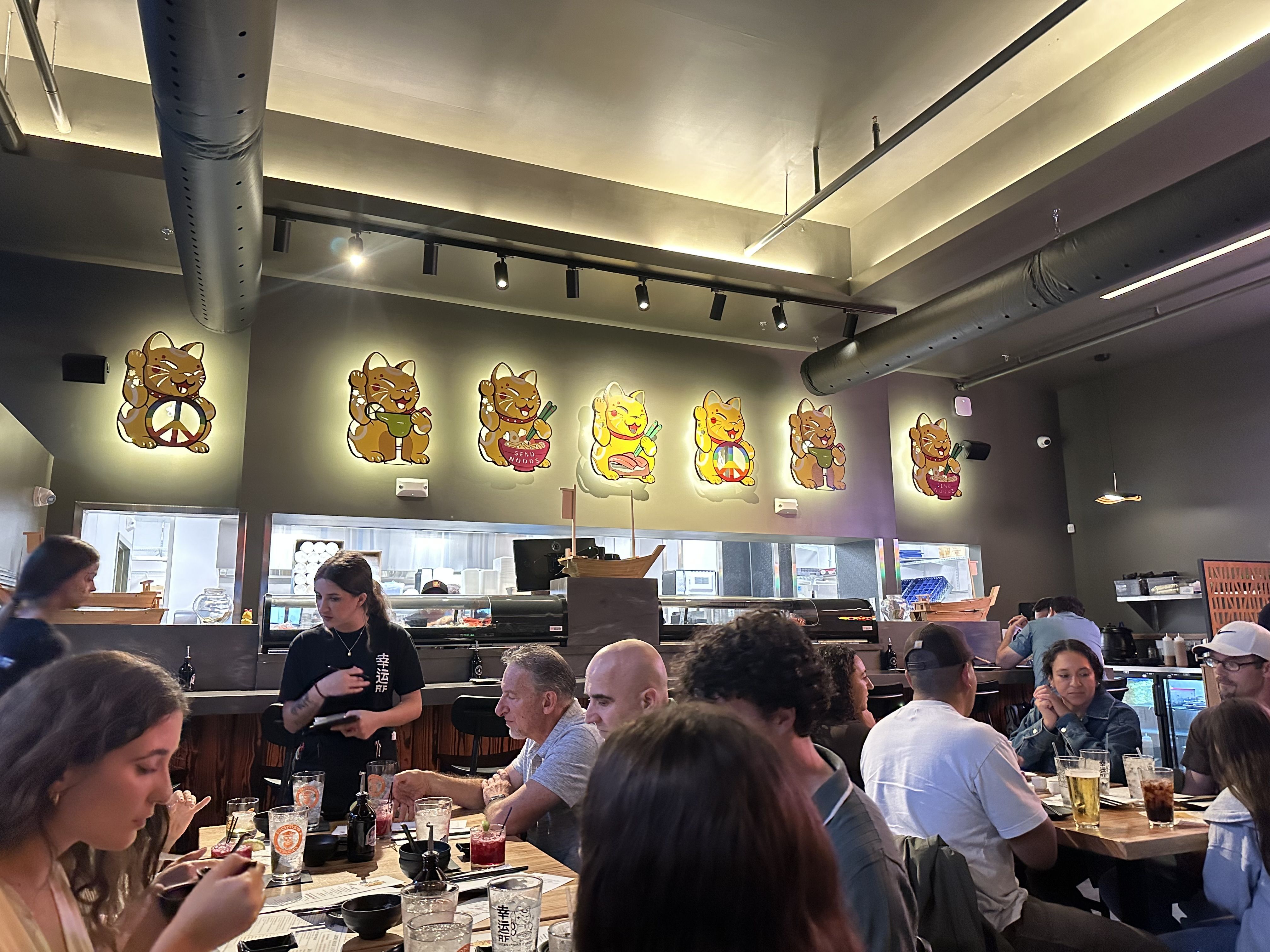 Busy restaurant interior with exposed ceiling ducts and track lighting. A row of colorful Maneki-neko cat wall art waves from the back wall as diners sit at tables and staff move about.
