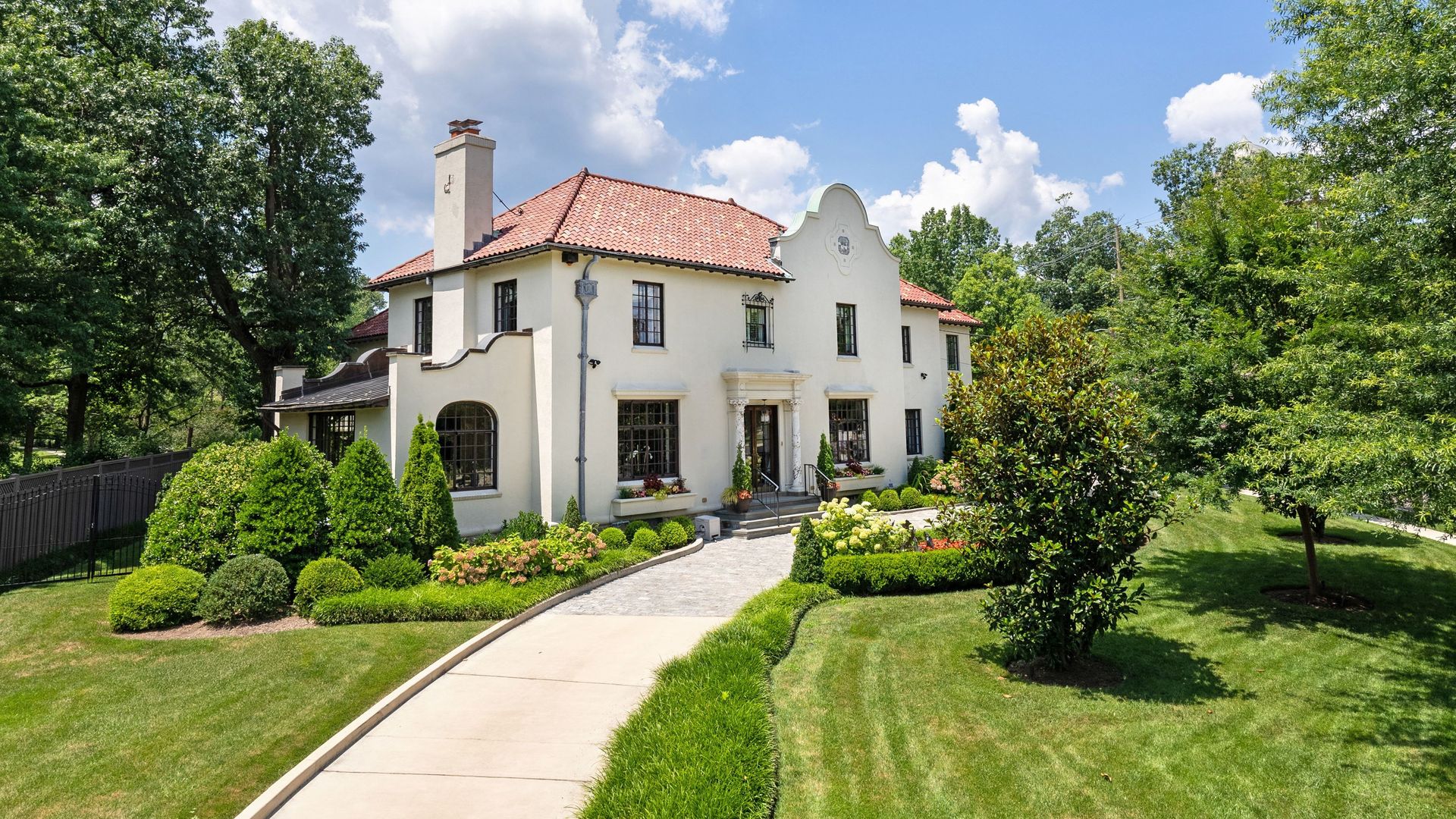 A white stucco two-story house with a red tile roof, a decorative curved front, arched windows, and a manicured lawn with trimmed hedges and colorful flowers; a curved walkway leads to the entrance.