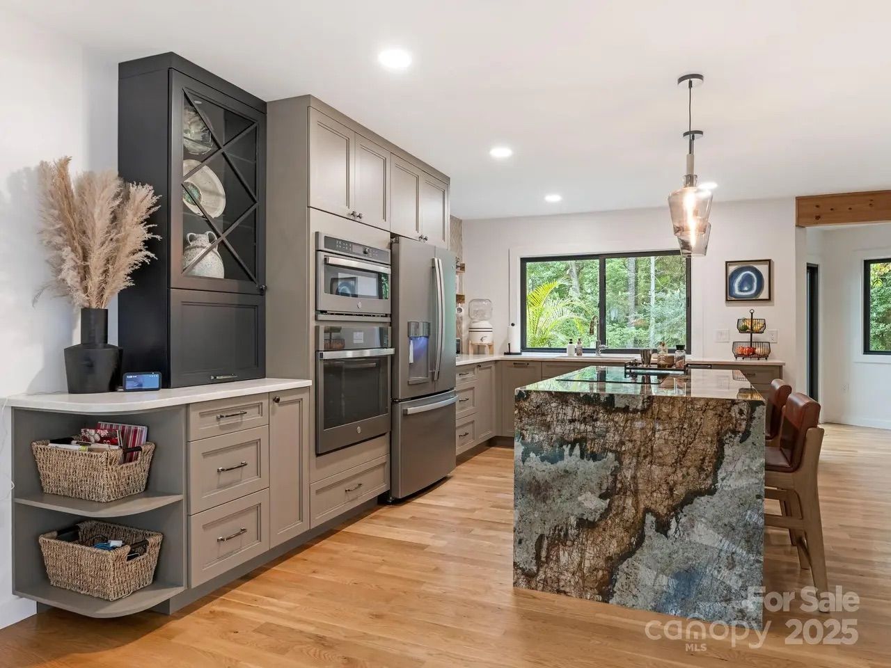 Modern kitchen with beige cabinets, stainless steel appliances, and a large island with a unique brown and blue stone finish, wooden floor, large window with forest view, and pendant light.
