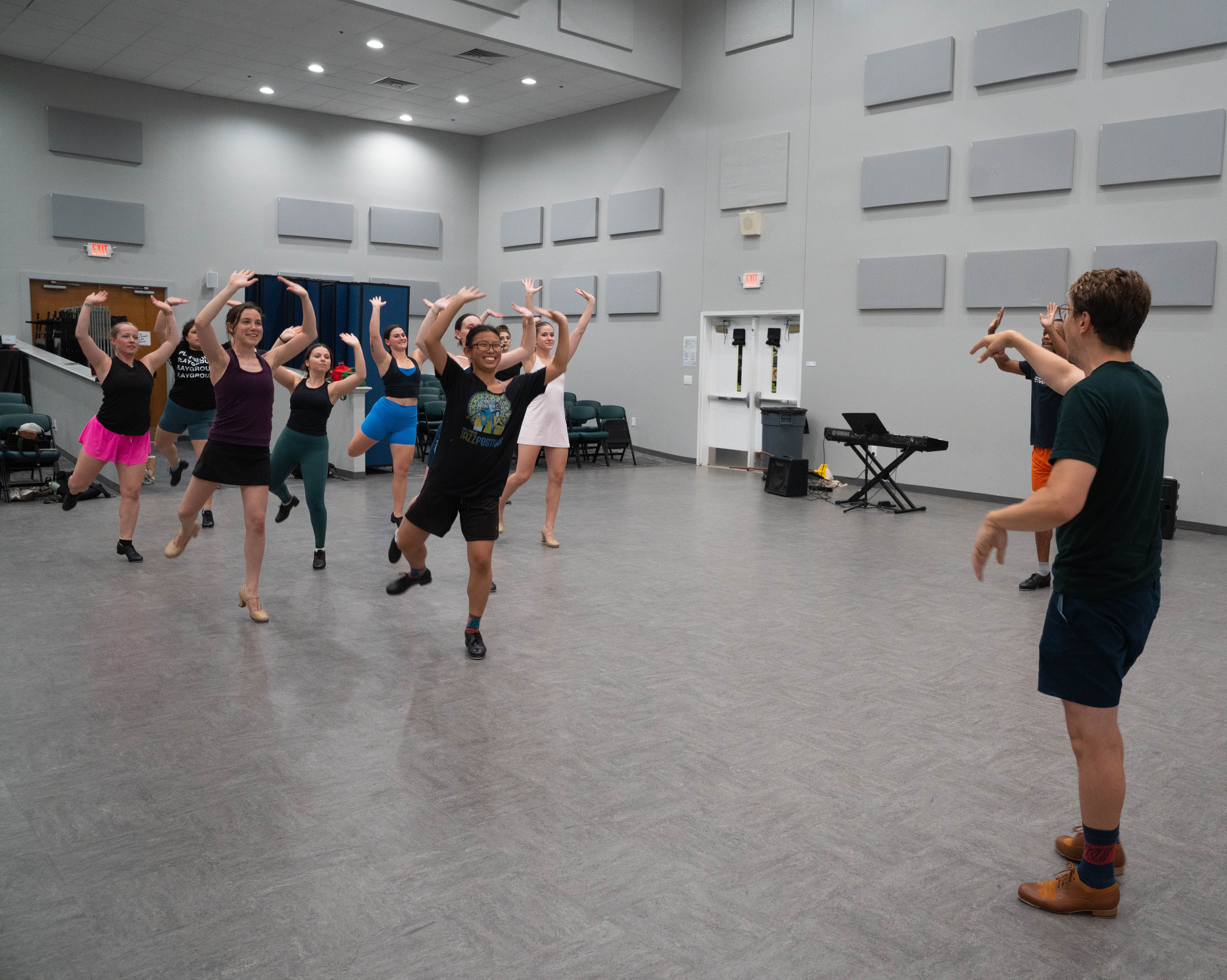 Group of people dancing in a large rehearsal room with grey walls and acoustic panels; front dancer raises arms and smiles as others follow, a man at a keyboard watches.